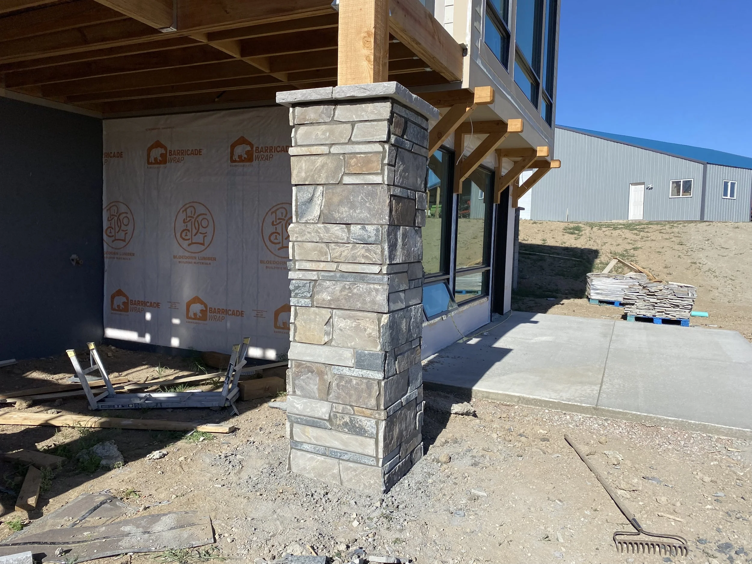 Construction site of a building with a stone pillar, large window, concrete sidewalk, and construction materials and equipment visible.