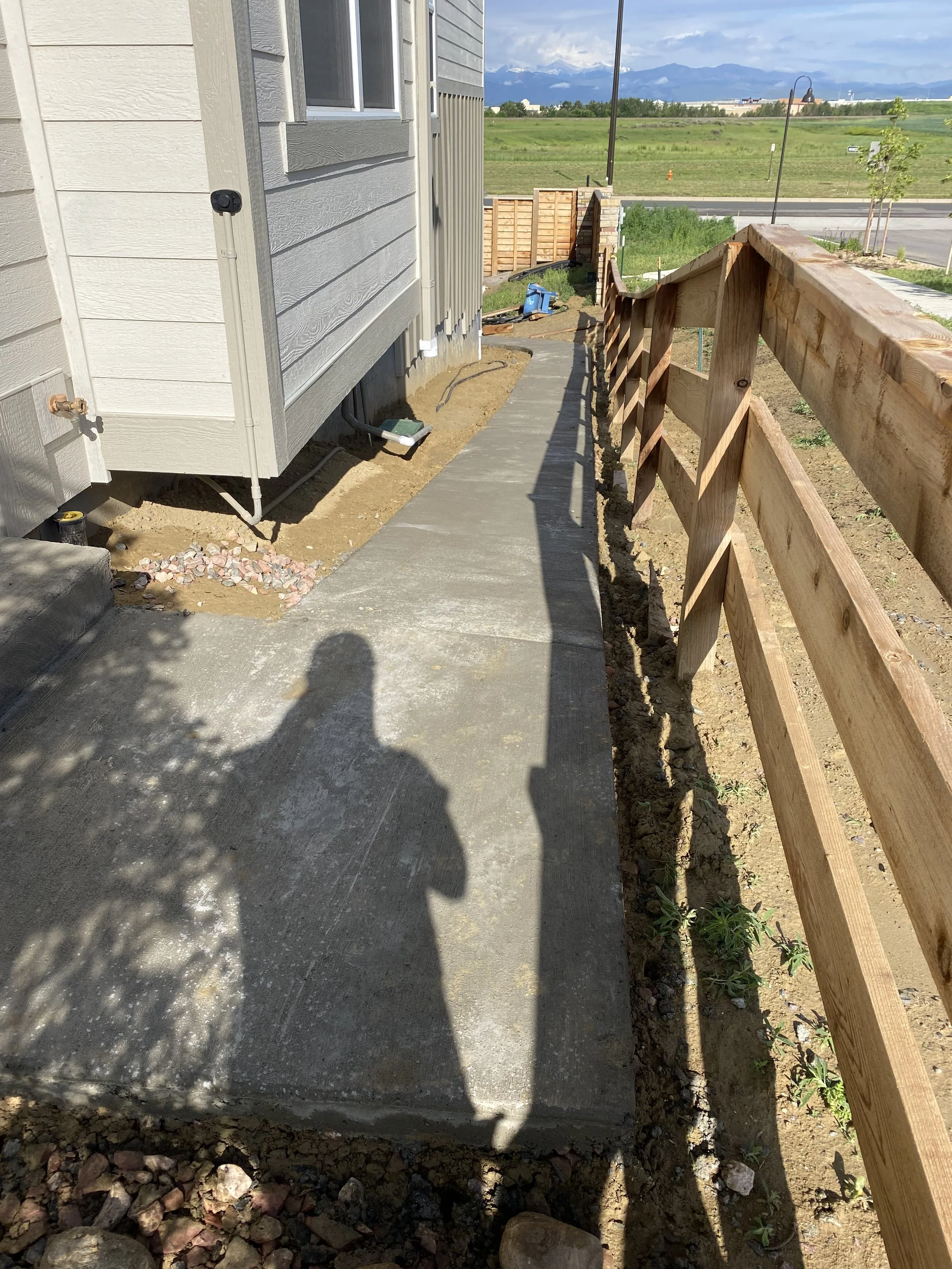 A newly poured concrete walkway next to a house with gray siding, bordered by a wooden fence and bordered on the other side by dirt and small plants, with shadows of two people cast on the concrete.