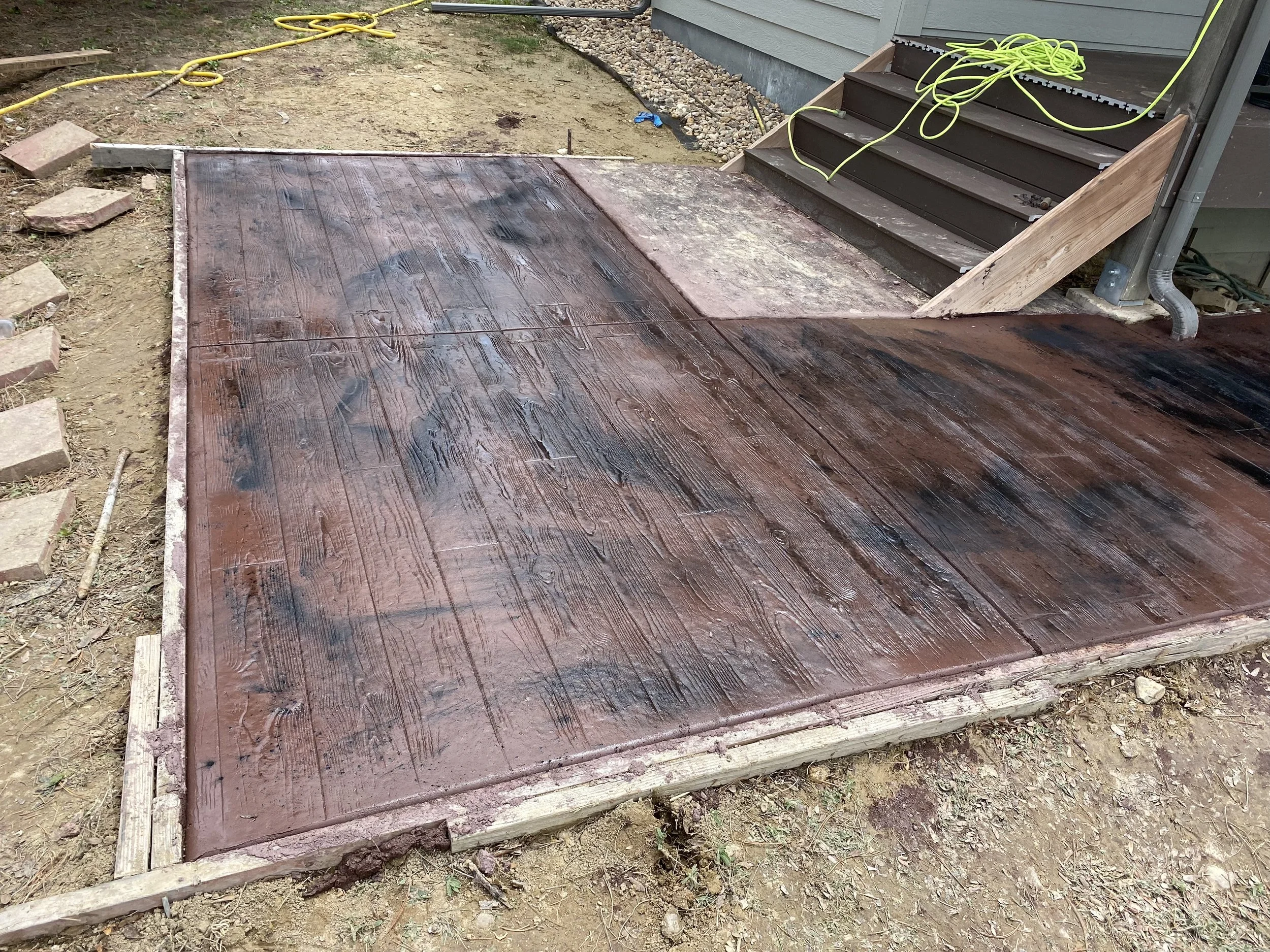 A new wooden porch being stained, with the stain still wet and partially applied on the planks, outside a house with steps leading up to a door.