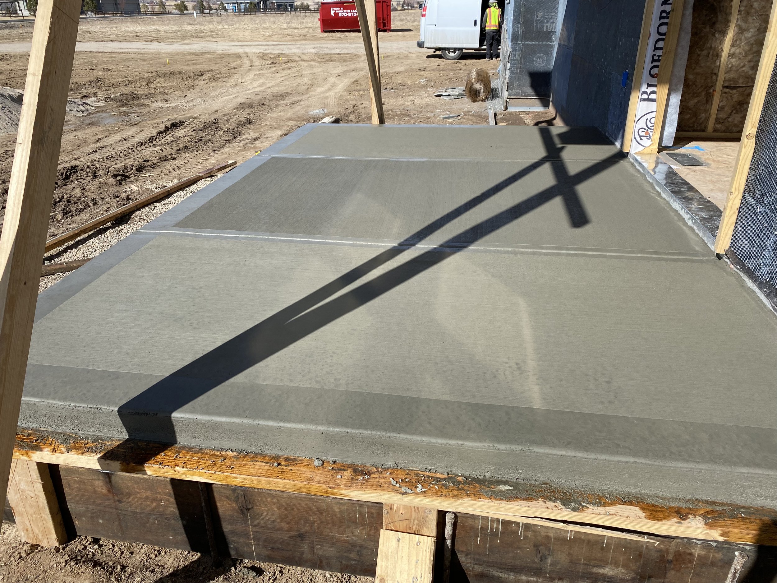 Freshly poured concrete slab on a construction site, with wooden formwork and a shadow of a cross-shaped structure cast on it.