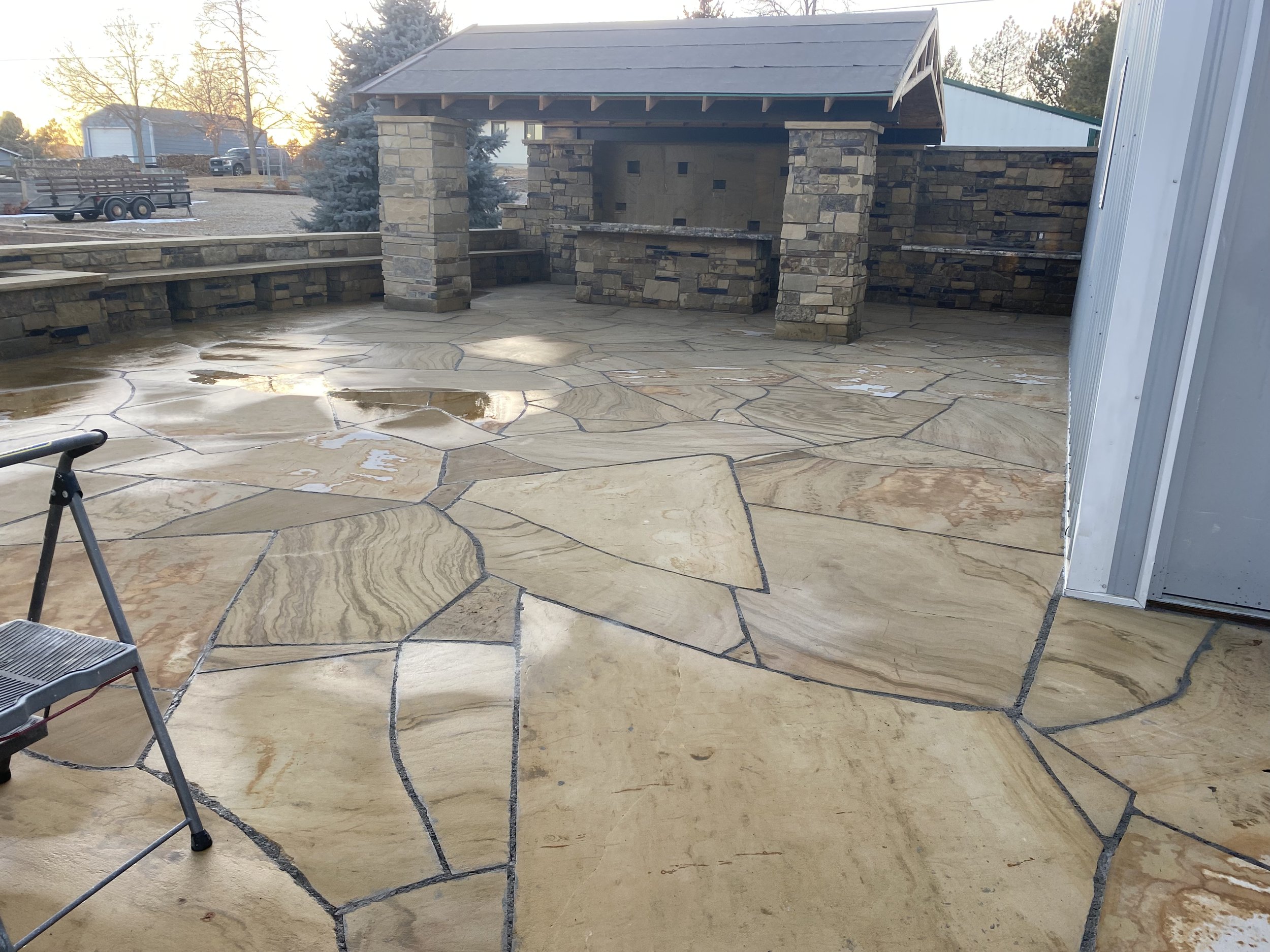 A stone patio with irregularly shaped beige and tan flagstones, some with water puddles. In the background, there's a stone wall with a covered outdoor cooking area or bar with supporting stone columns and a wooden roof, set against a sunset sky and trees.