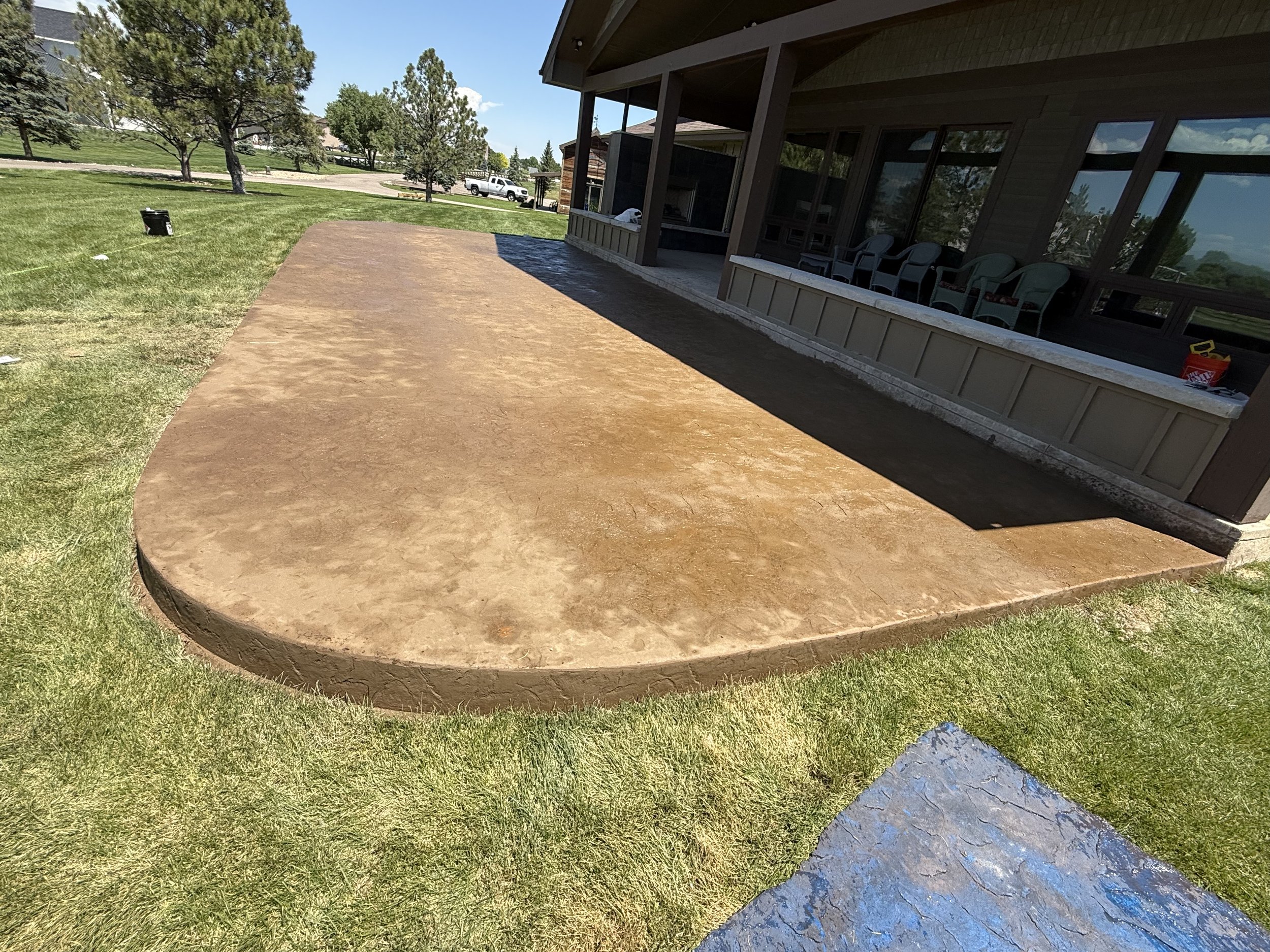 A freshly poured concrete patio with rounded edges next to a house, surrounded by green grass and a few trees in the background.