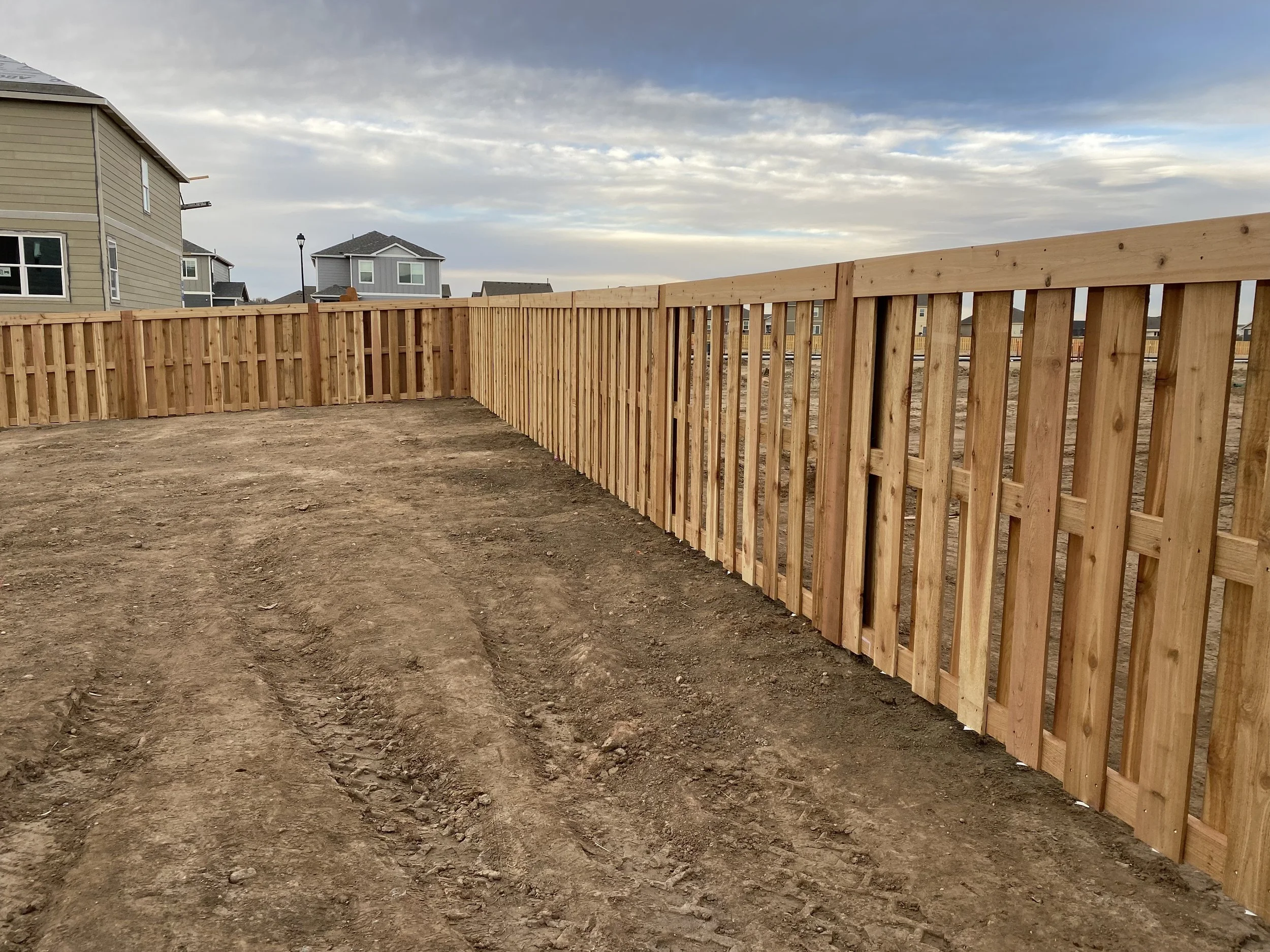 A newly built wooden fence surrounded by dirt on a construction site with suburban houses in the background under a cloudy sky.