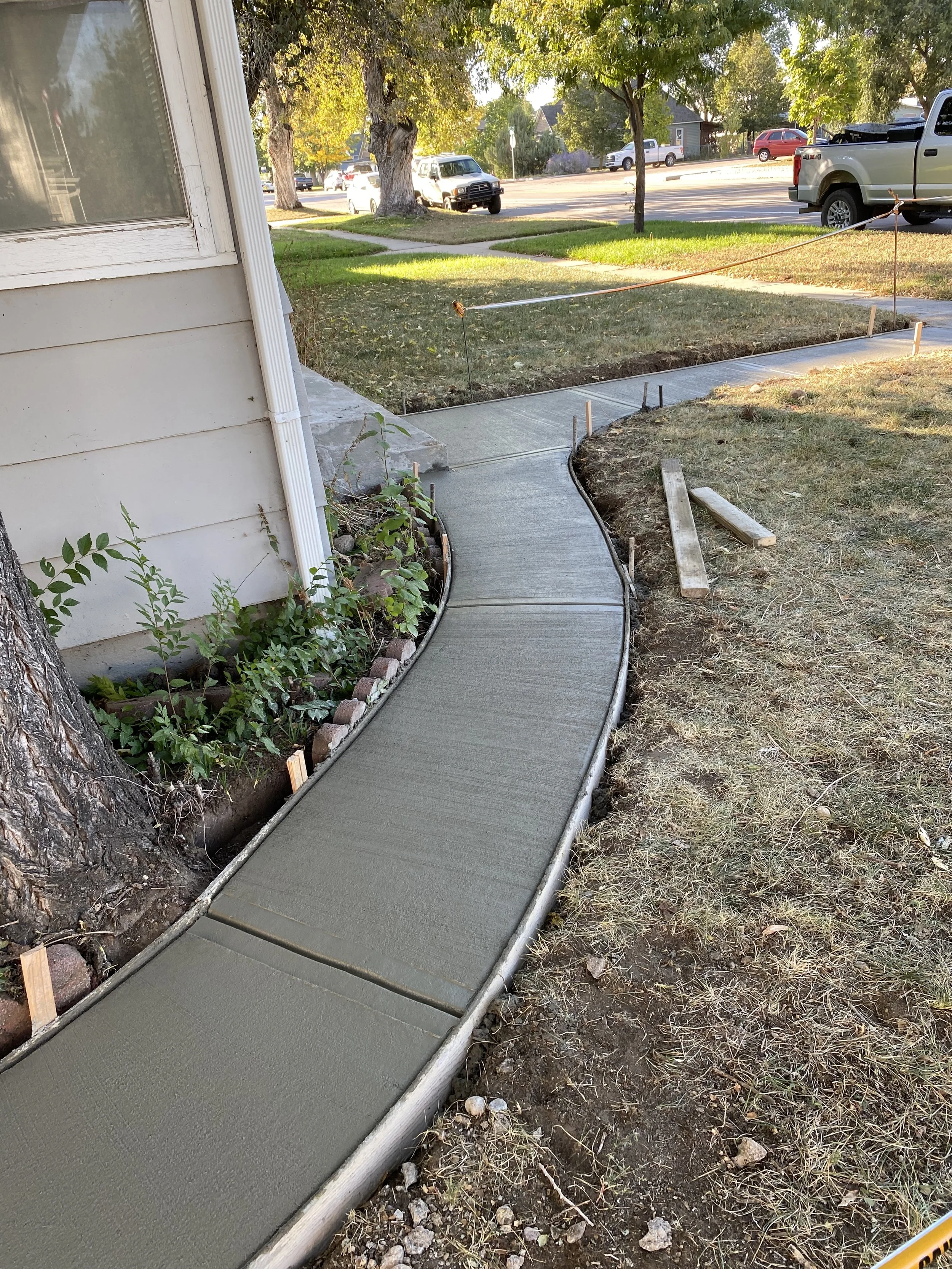 Newly poured concrete walkway with curving design in front of a house, bordered by a garden bed and trees, with construction markers and wooden stakes in place.