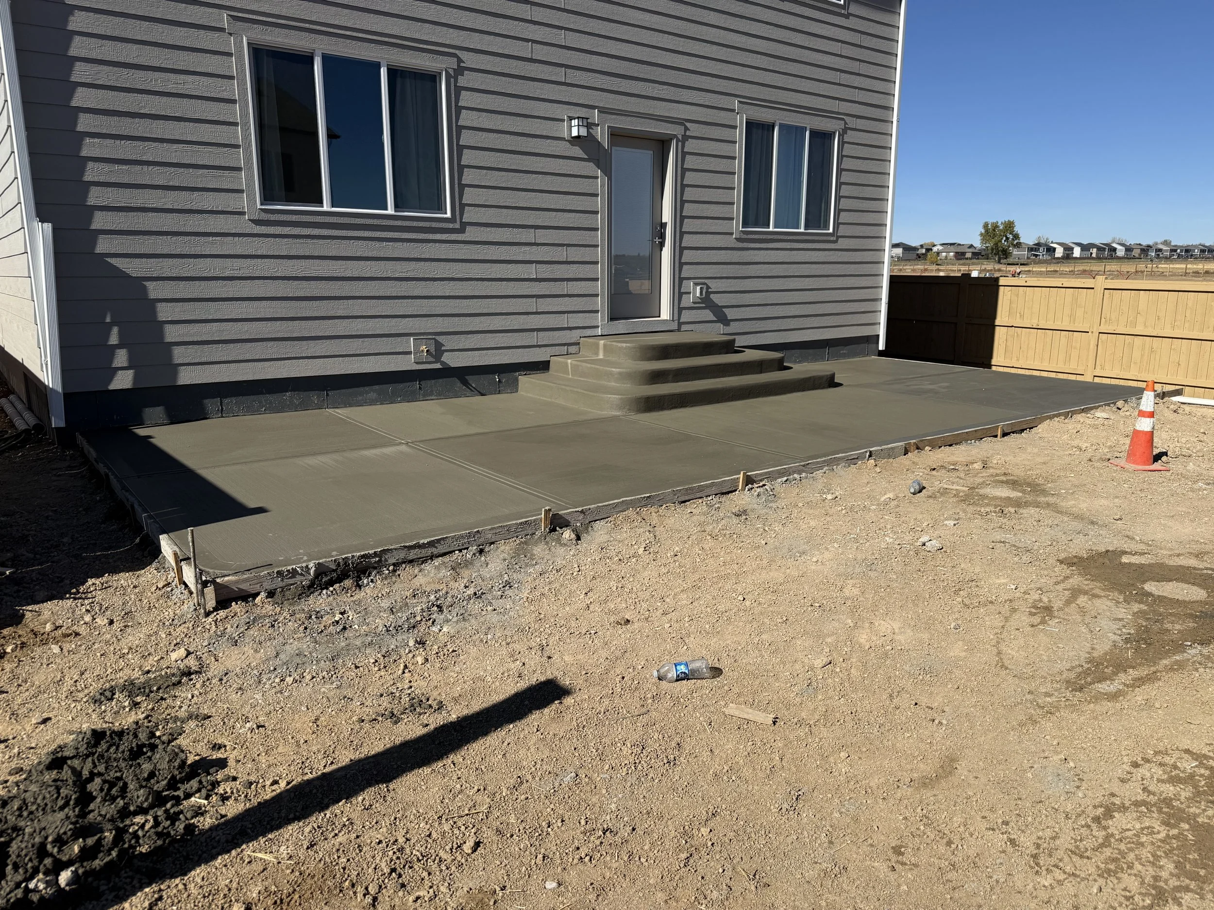 Concrete patio being poured in front of a house with gray siding, with steps leading to a back door. Construction cones and uneven dirt surface with a plastic water bottle in the yard.