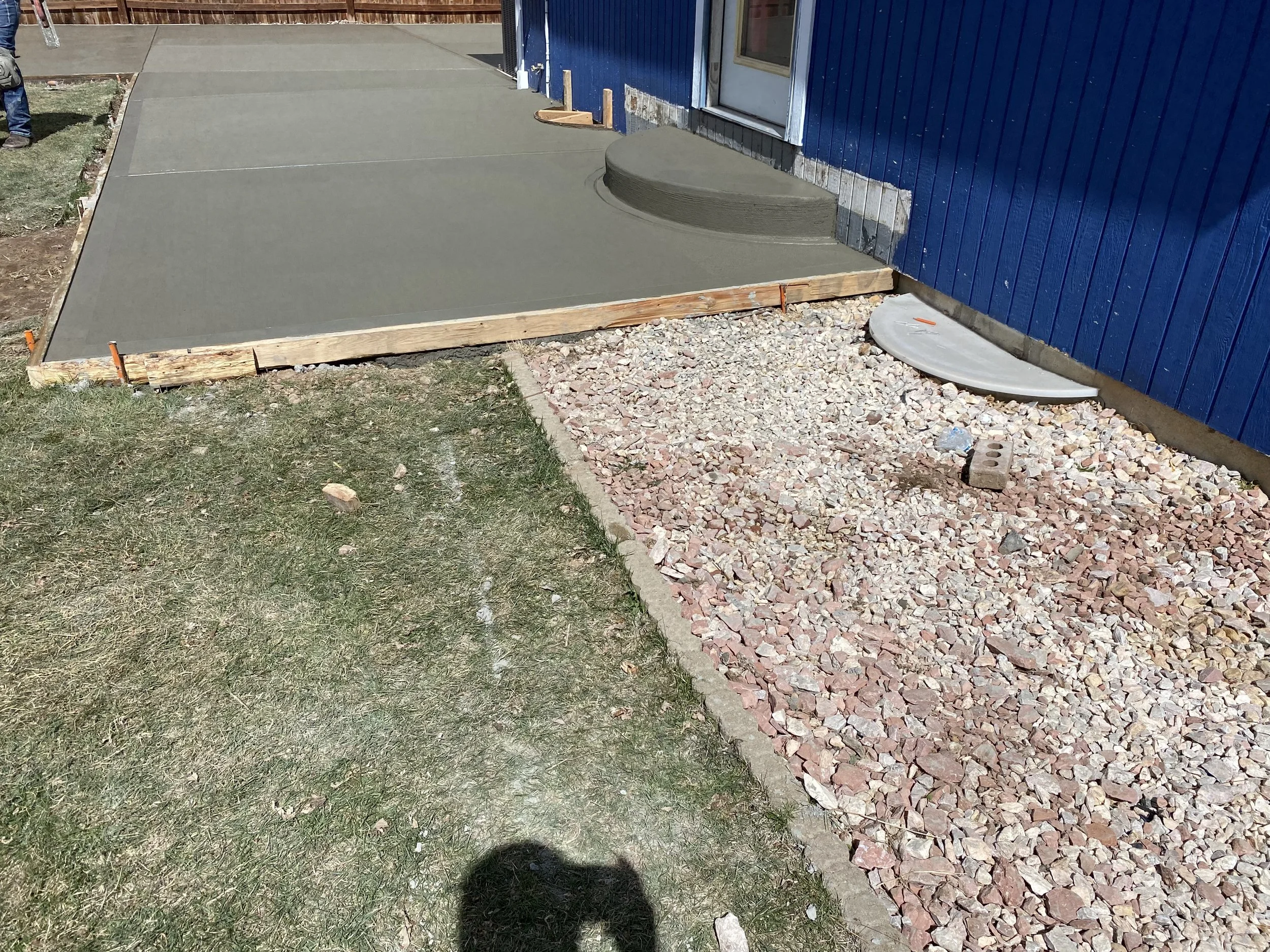 Newly poured concrete sidewalk with wooden forms, adjacent to a blue building with gravel and stepping stones, and a small grassy area.