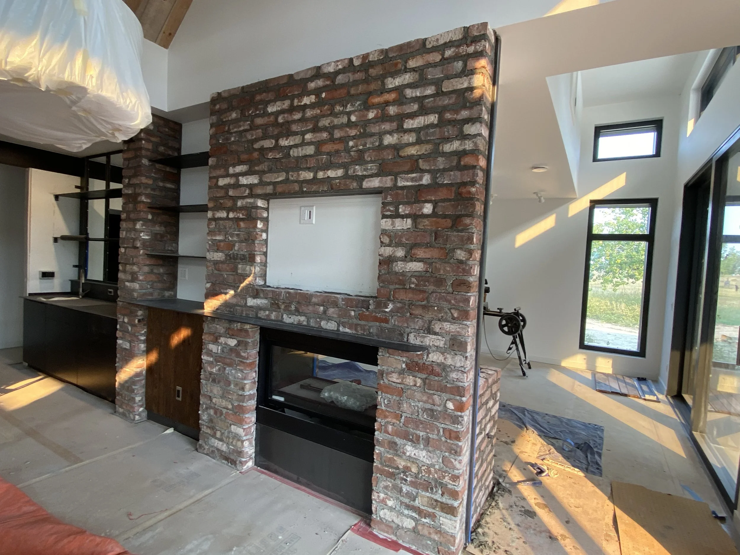 Interior view of a modern home with a brick partition wall featuring a fireplace and open shelves. Sunlight streams through large windows, illuminating the space which appears to be under construction or renovation.