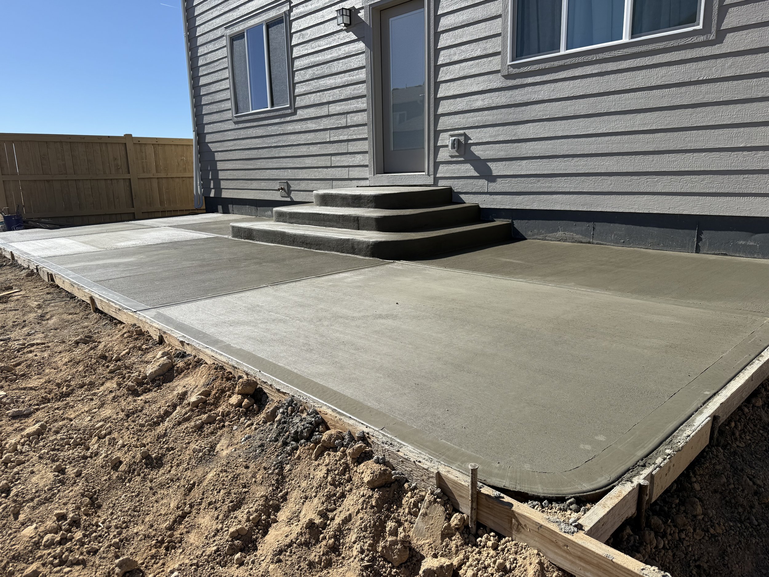 Newly poured concrete patio with four steps leading to a house door, in a backyard with a wooden fence and dirt surrounding the patio.