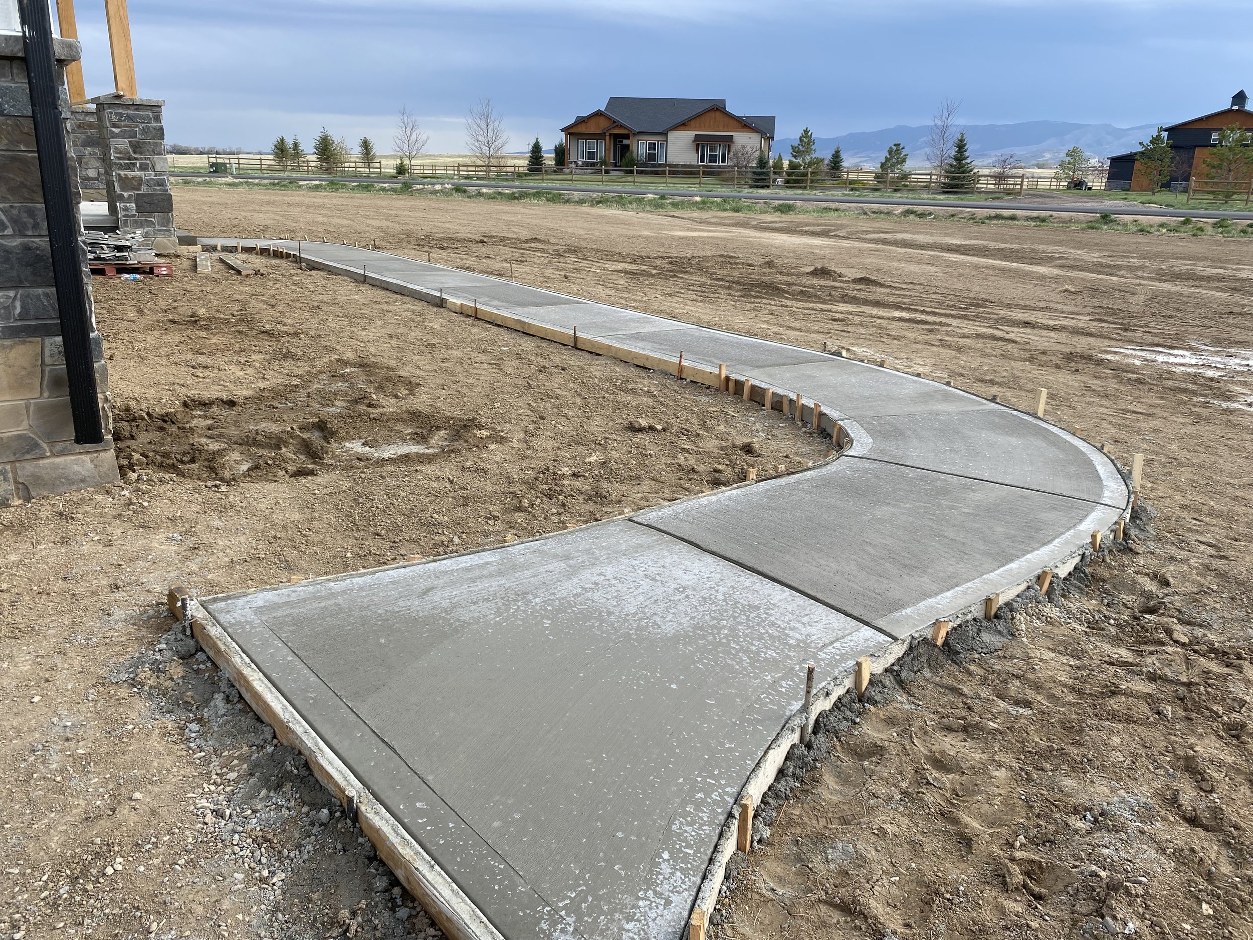 Newly poured concrete sidewalk with a curved path on a construction site, with wooden forms in place and dirt surrounding the area. In the background, there are residential houses with fencing and mountains in the distance.