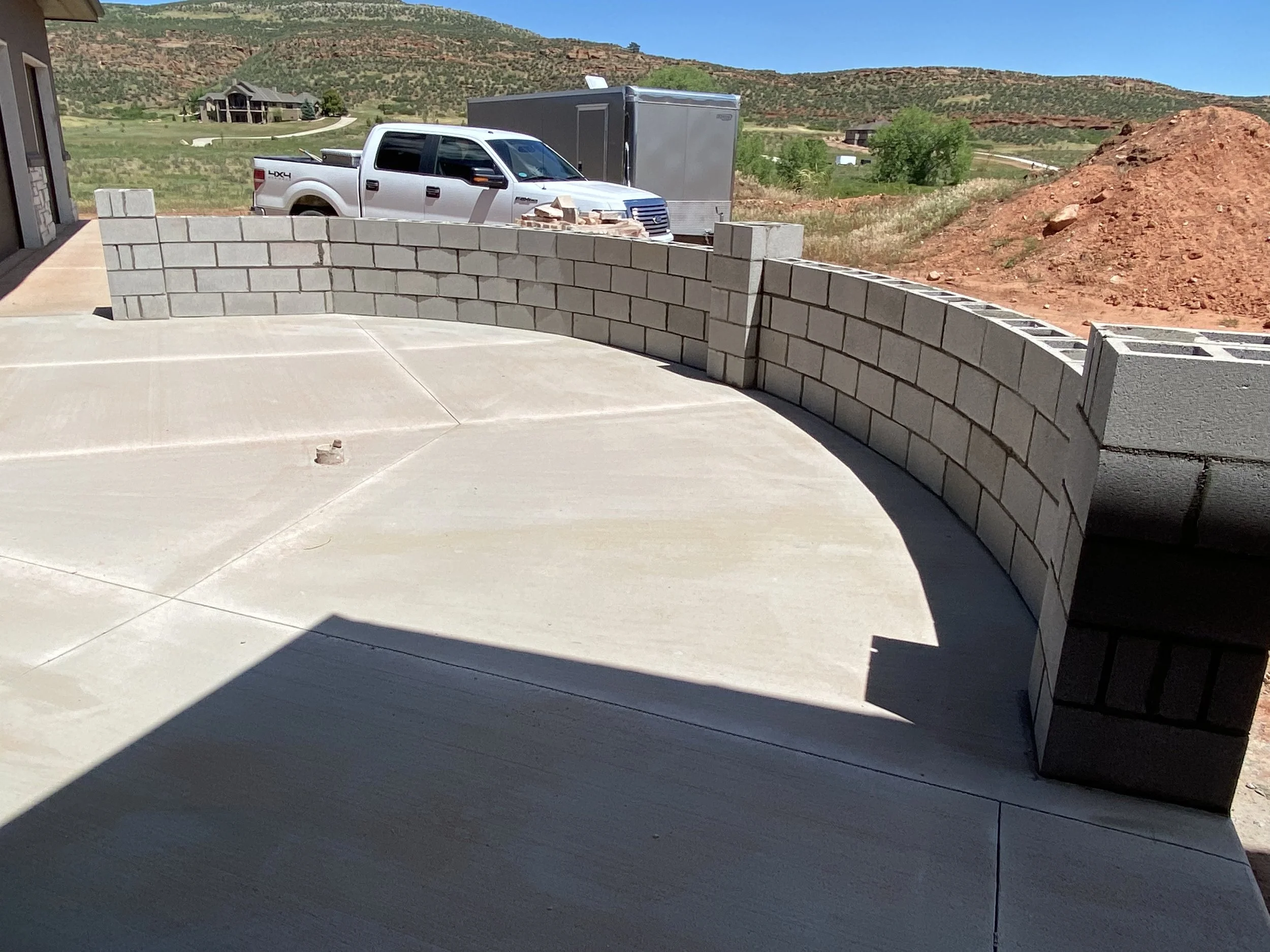 A curved brick wall being built on a concrete patio with a white pickup truck and a gray trailer in the background, set in a desert landscape with hills and a house in the distance.