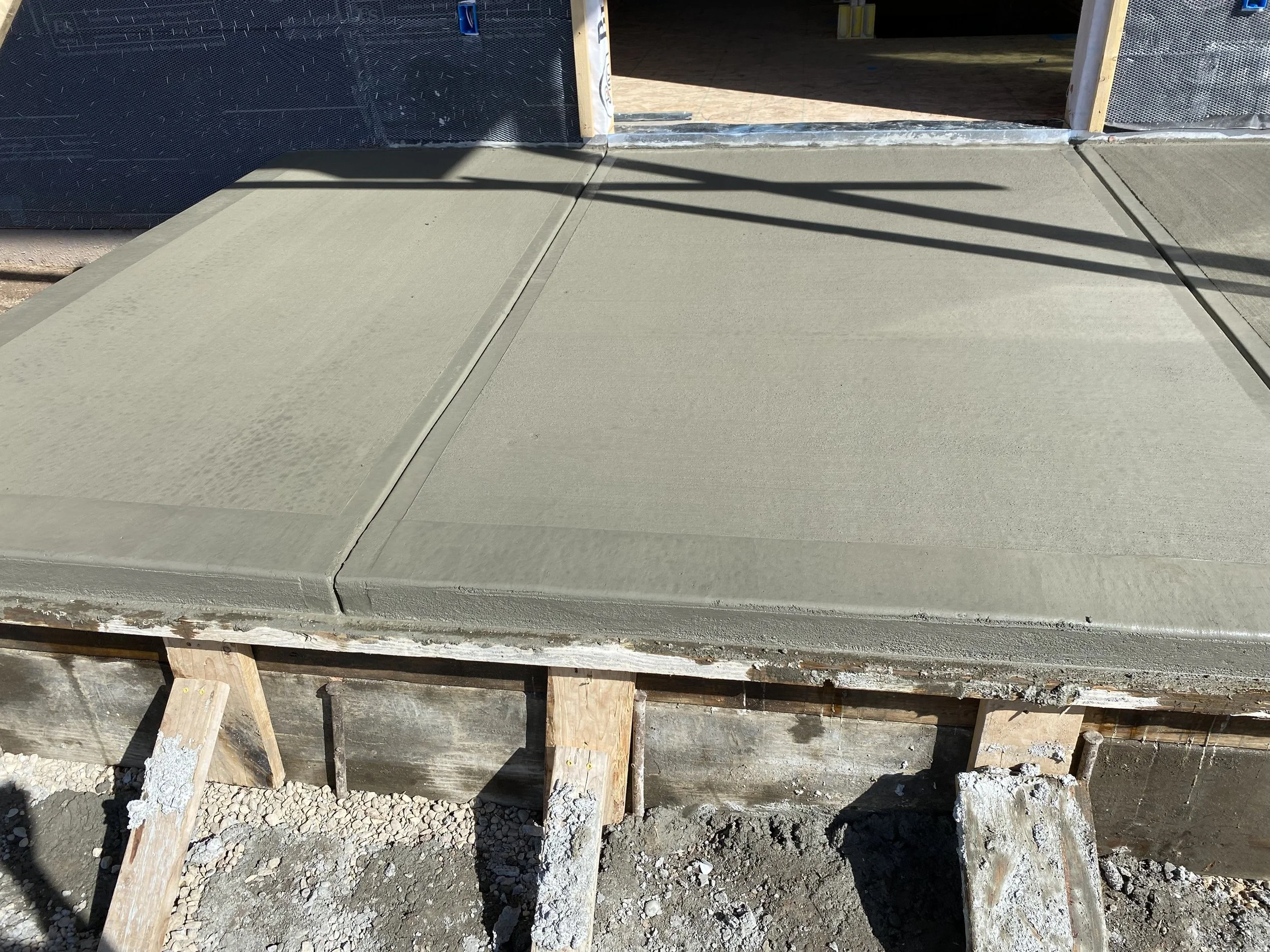 Freshly poured concrete slab on a building foundation with wooden supports underneath, partially in shadow.