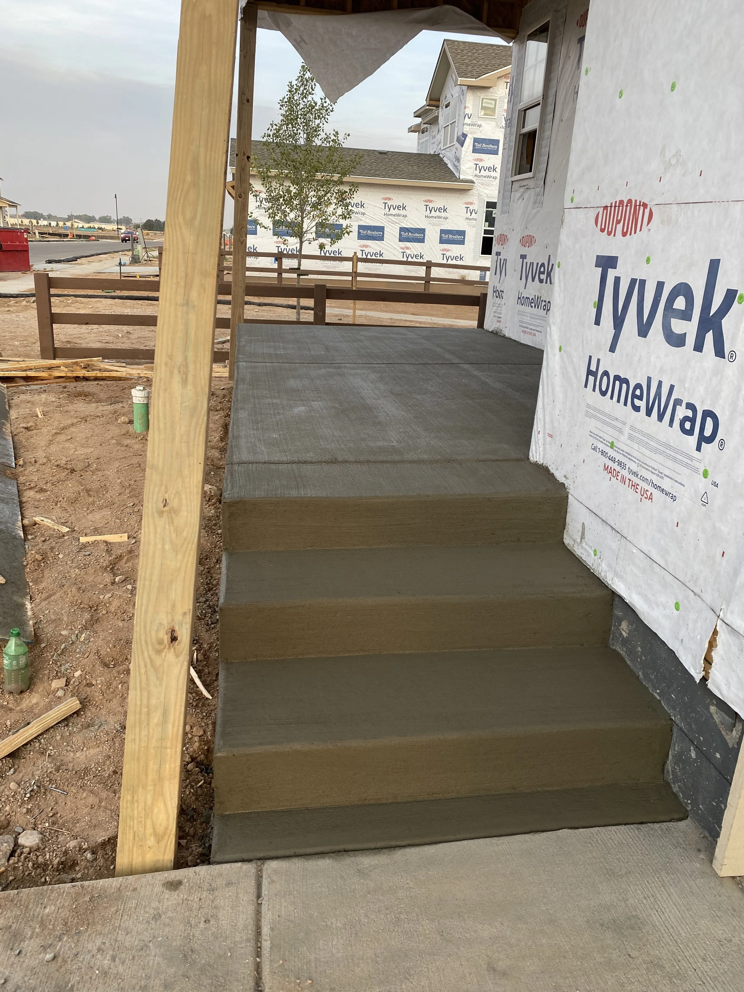 Construction site with newly built concrete stairs leading to an elevated porch. The house exterior is wrapped in white Tyvek HomeWrap.