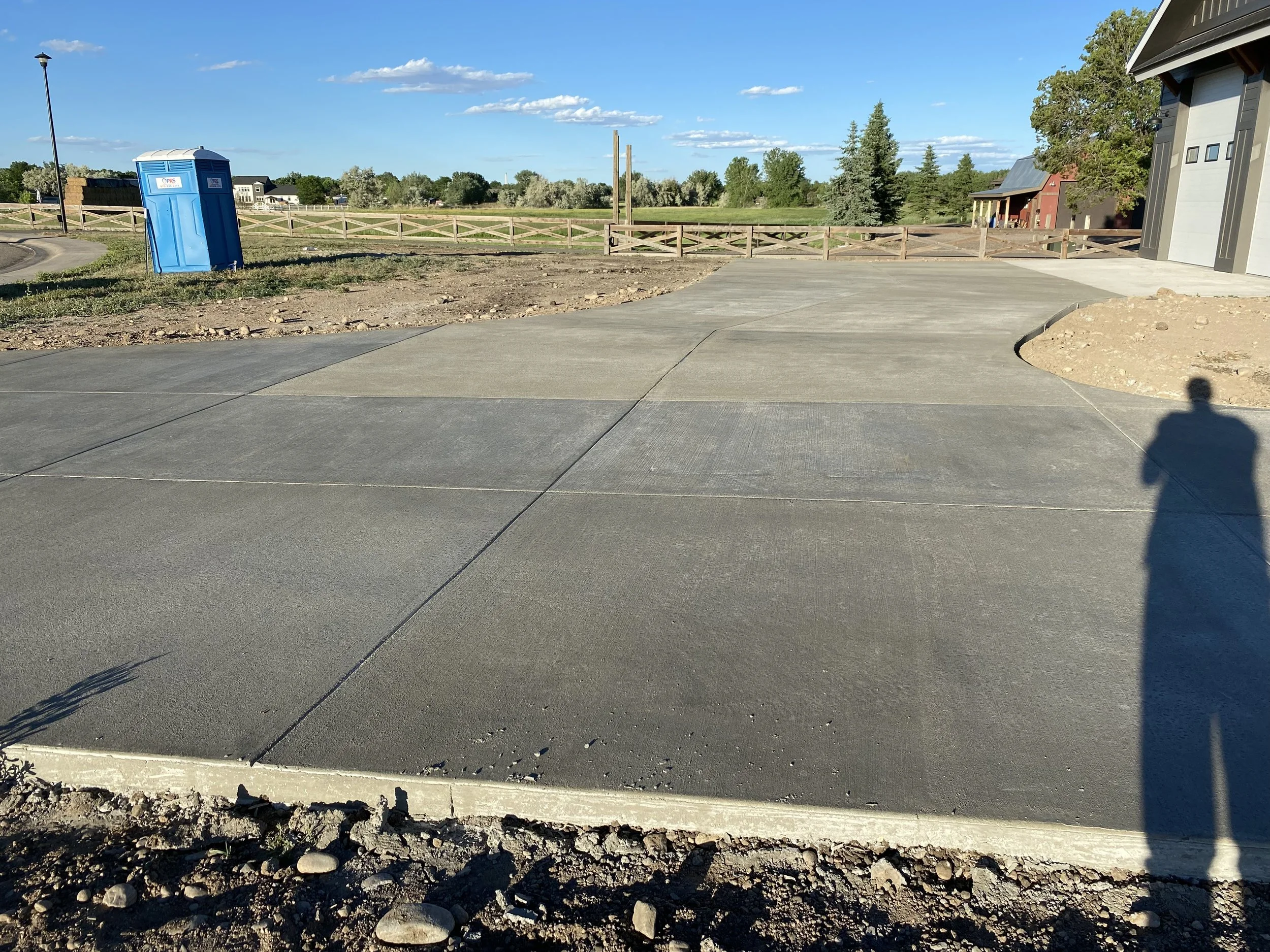 Newly poured concrete driveway with visible expansion joints, bordered by dirt and a concrete curb, in a semi-rural area with a blue portable toilet, trees, a red building, and a garage door, on a sunny day with a person's shadow visible.