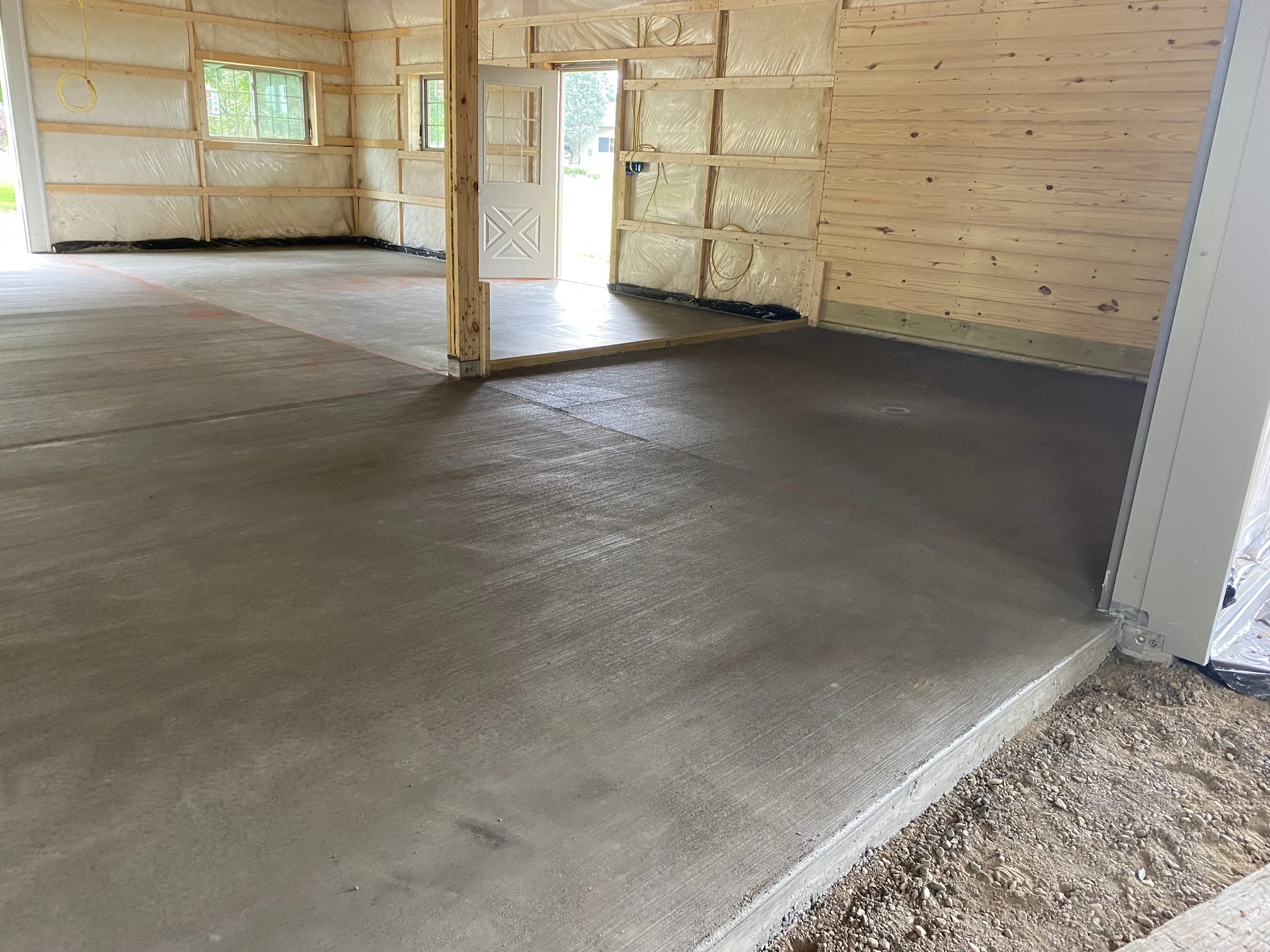 Interior of a house under construction with a newly poured concrete floor, unfinished wooden walls, and a white door with windows leading outside.