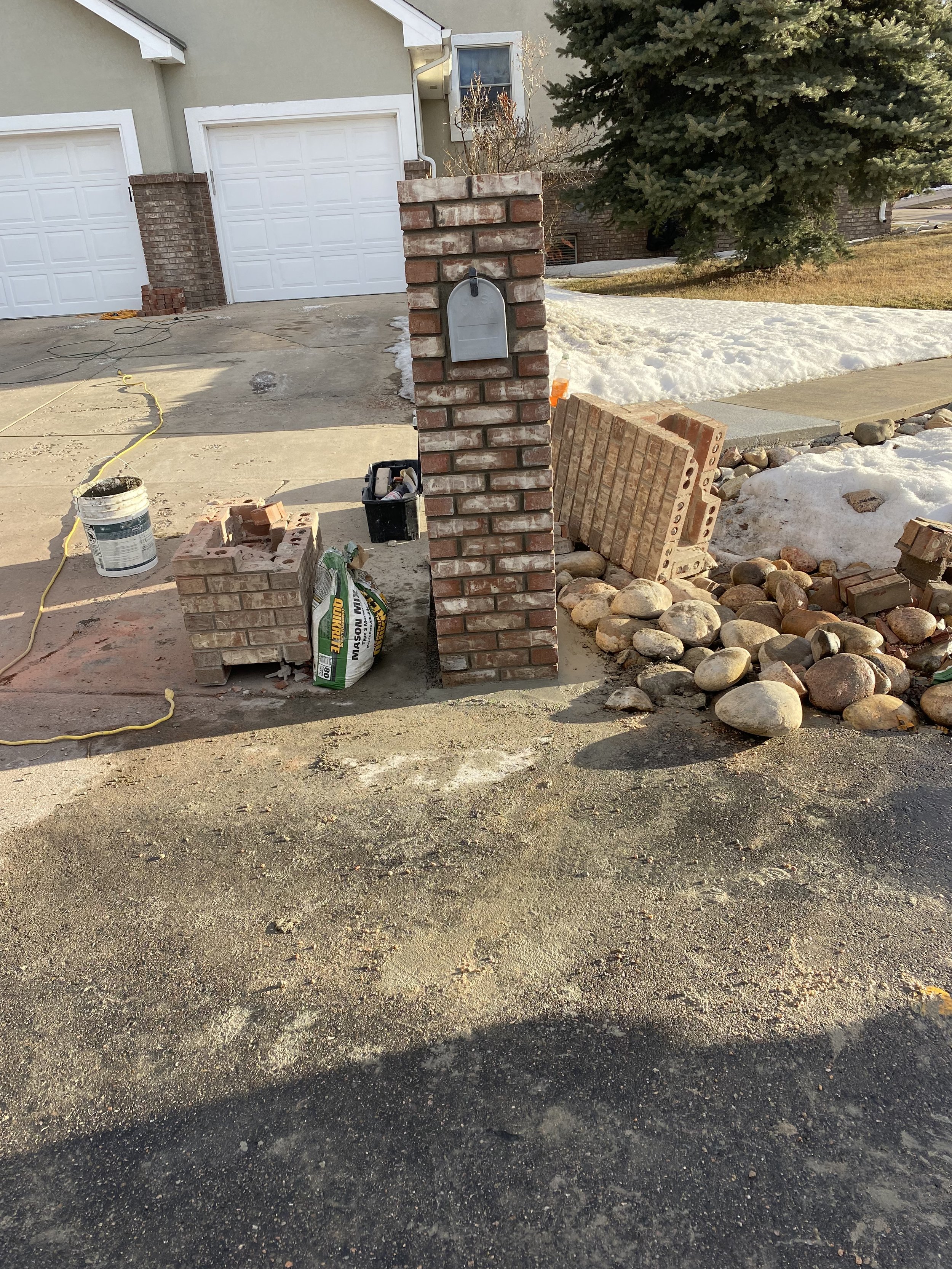 Construction scene in front of a house showing a brick pillar with a mailbox, construction materials including bricks and a bag of mortar, rocks, and a partially snow-covered yard/background.