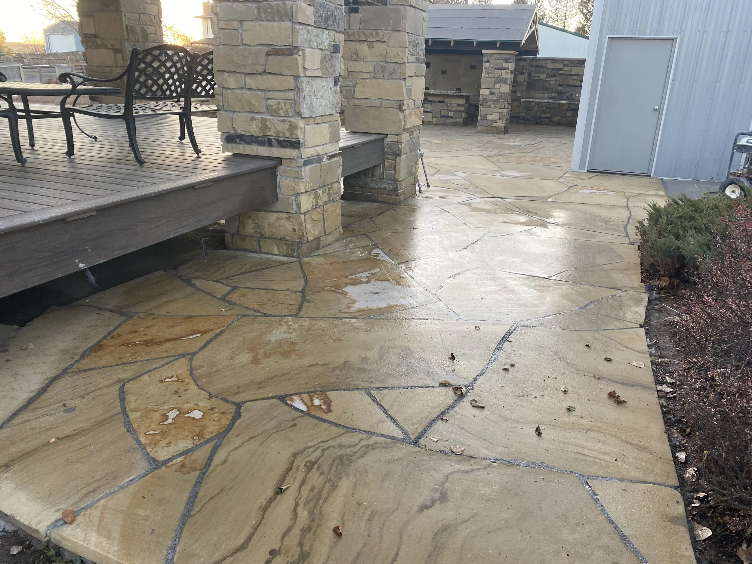 Stone and concrete patio with a wooden deck and chairs, adjacent to a stone covered outdoor kitchen and a metal shed.
