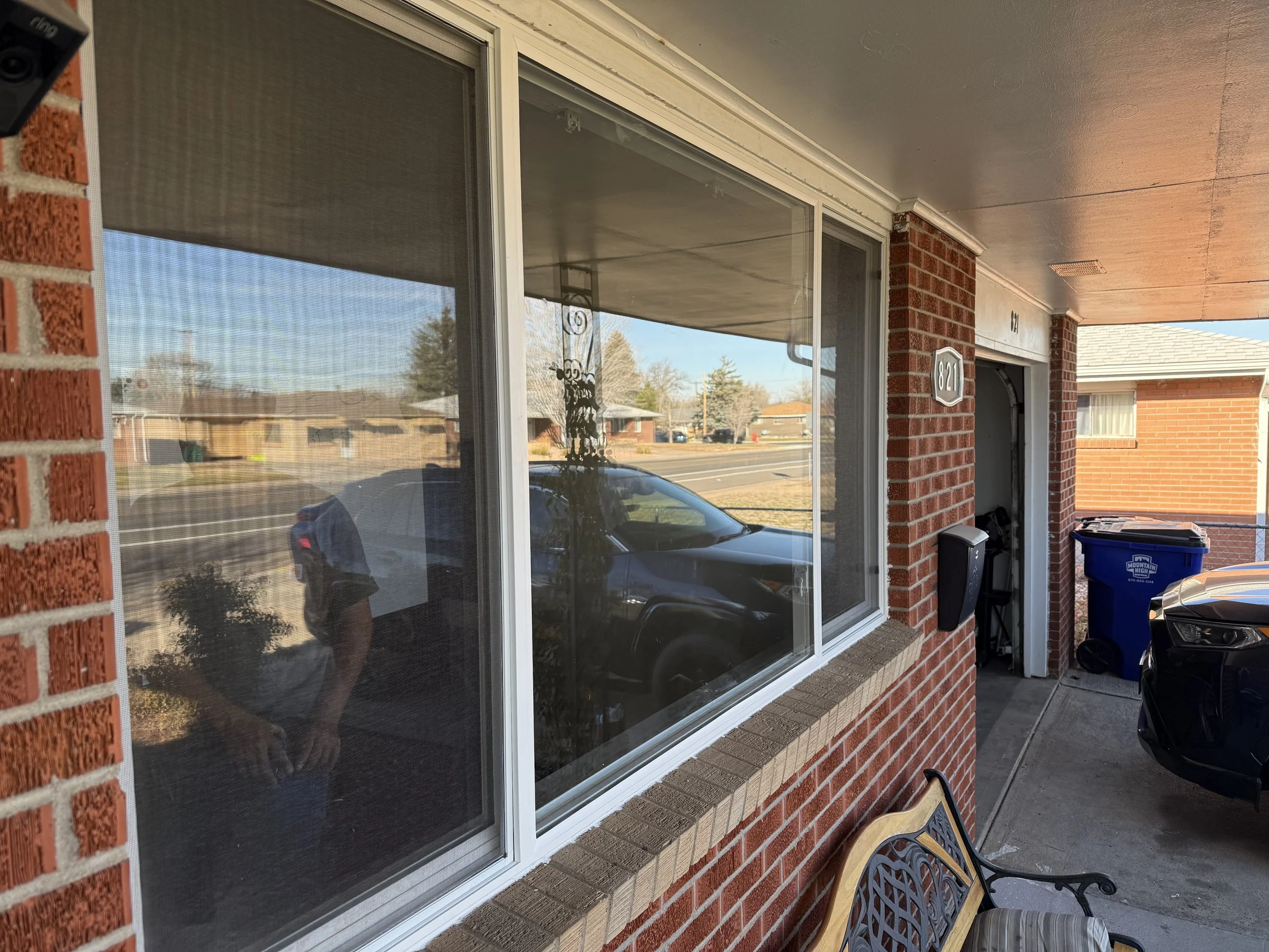 Front porch of a brick house with large window, a black vehicle parked nearby, trash bins, and a bench.