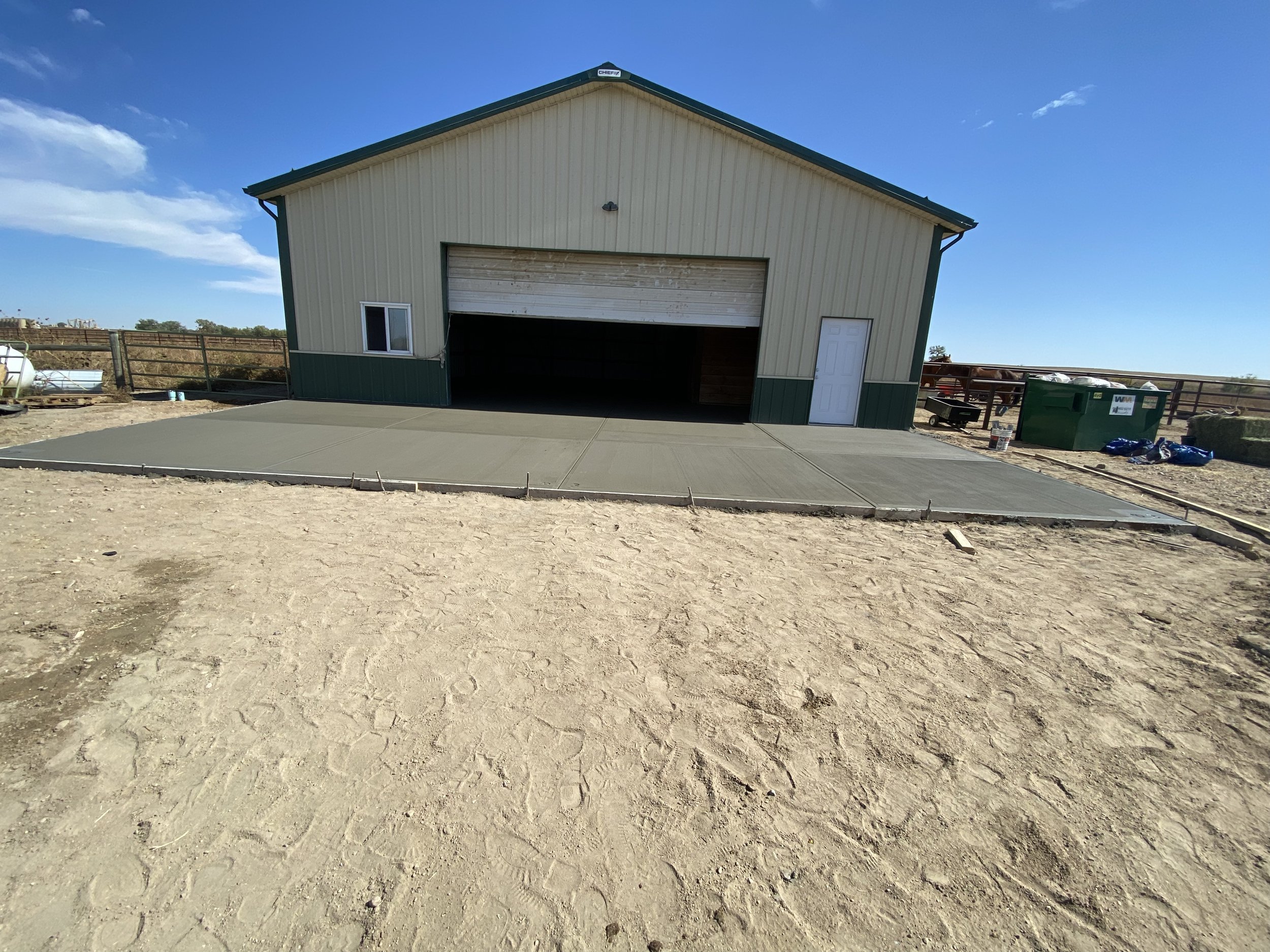 A large metal building with a roll-up garage door and a side door, situated on a flat concrete slab, with dirt ground and a blue sky in the background.