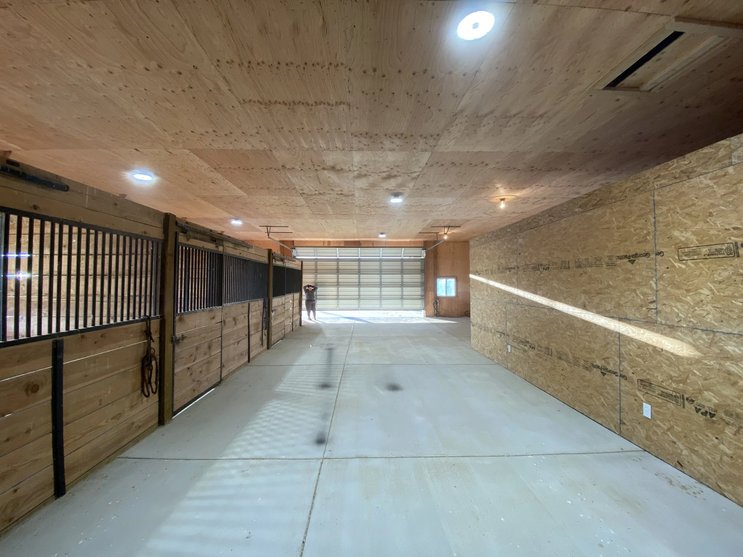 Interior of a garage or storage space with wooden walls and ceiling, concrete floor, and garage door at the far end. There are multiple ceiling lights, some turned on, and a person standing near the garage door.