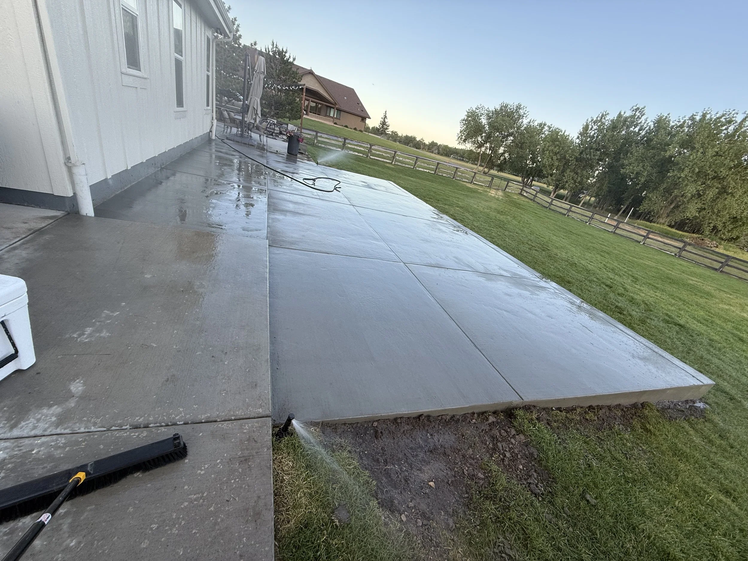 A concrete patio being washed with a pressure washer, with water spraying onto the surface near a house, surrounded by a grassy yard, trees, and neighboring houses in the background.
