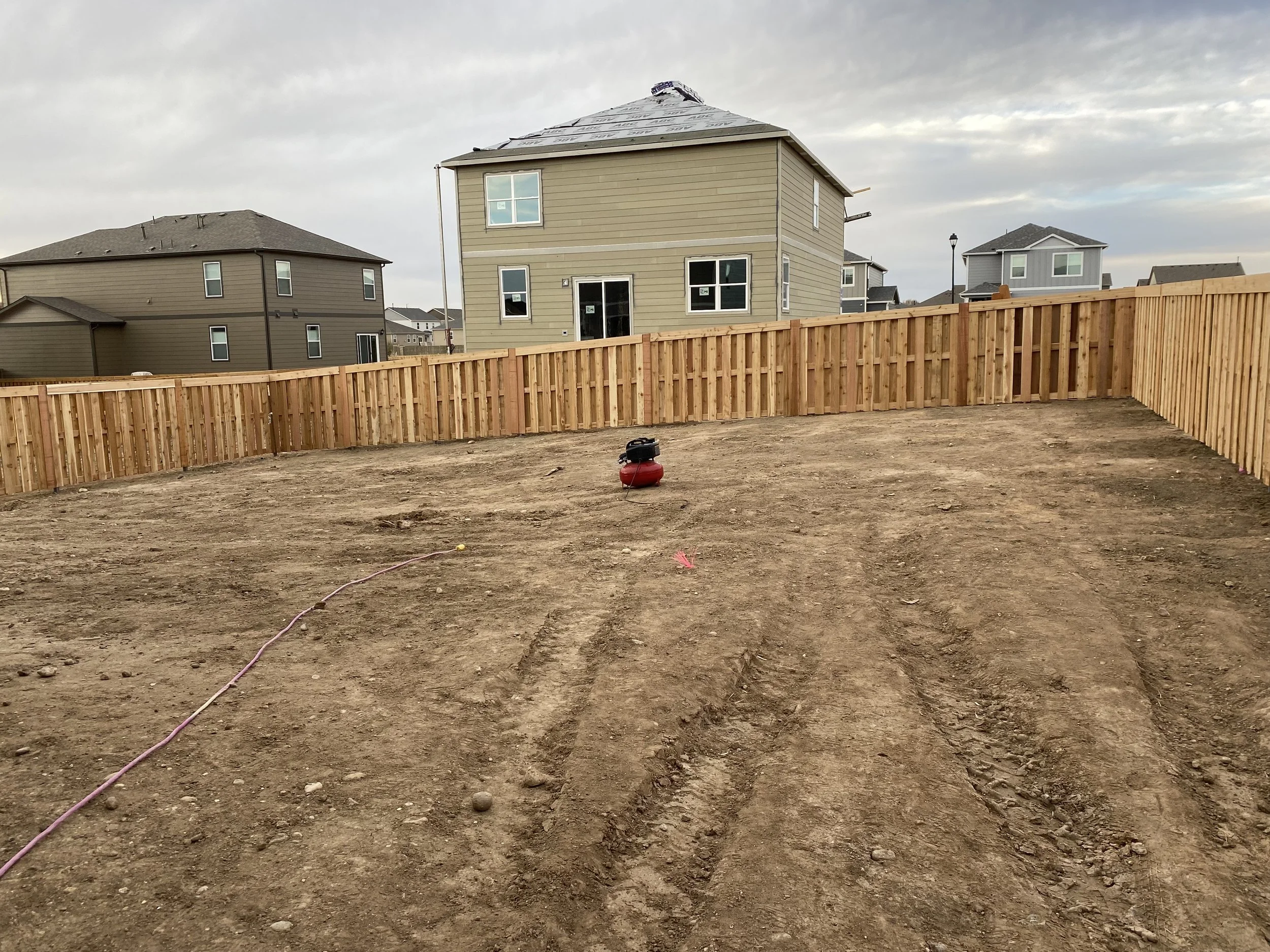 Empty backyard with dirt ground, a red and black outdoor power tool, and a new wooden privacy fence surrounding the yard. In the background, there are multi-story houses and a cloudy sky.