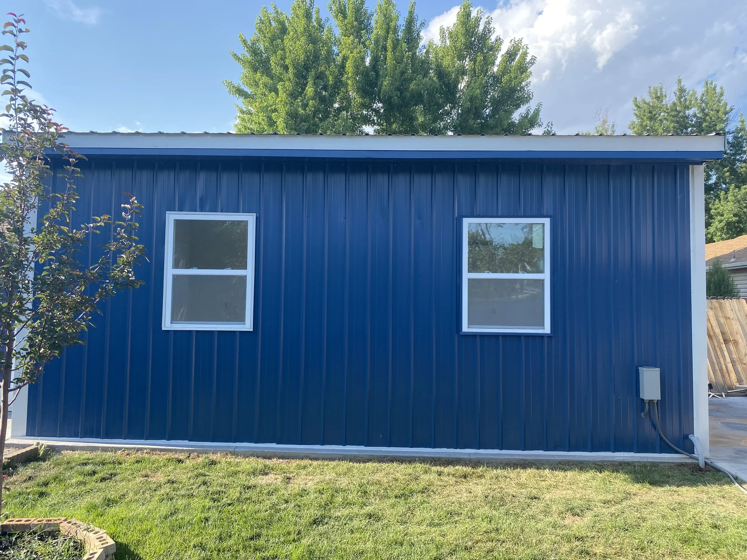 Back of a blue metal building with two windows, a tree on the left, and electrical box on the right, with a grassy yard in front.