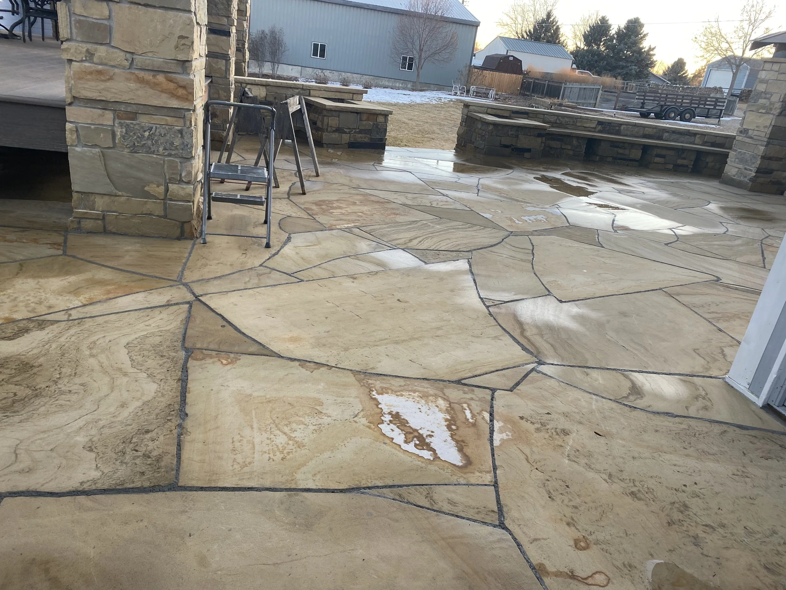 A concrete patio with polished, irregularly shaped sandstone tiles, some with a light coat of snow, surrounded by stone pillars and outdoor furniture, in a backyard at sunset.