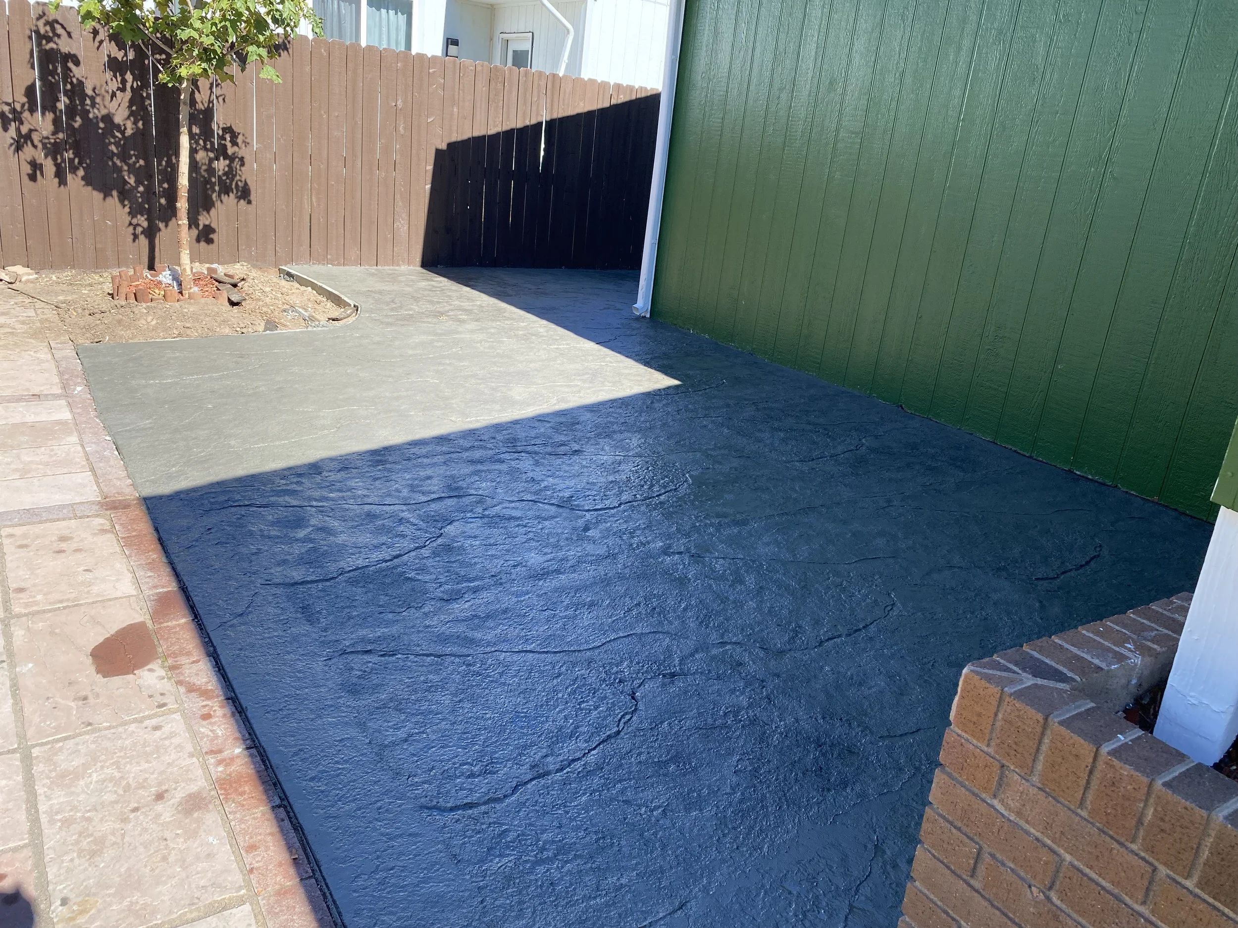 The image shows a backyard patio area with a new concrete slab that is freshly poured and has a textured, finished surface. There is a small tree planted in a circular brick-edged bed in the background, with a brown wooden fence and a green exterior wall of a building on the right side.