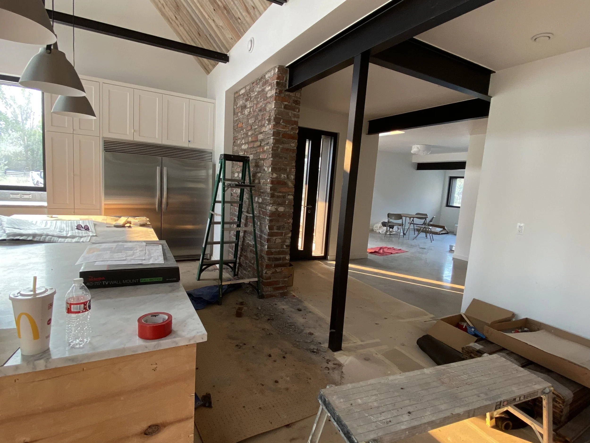 Interior of a house under renovation with exposed brick wall, black metal beams, a kitchen island with a McDonald's drink and water bottle, a ladder, and construction tools and materials scattered around.