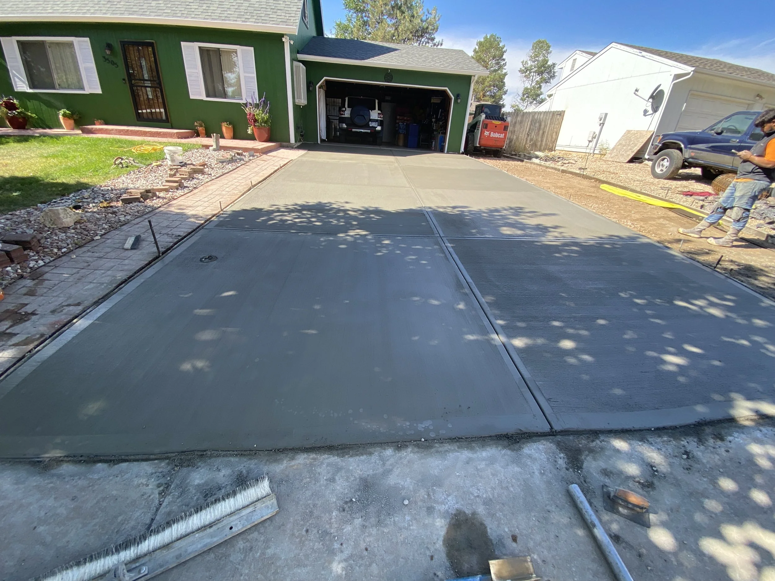 Newly poured concrete driveway in front of a green house, with construction tools and workers adding finishing touches.