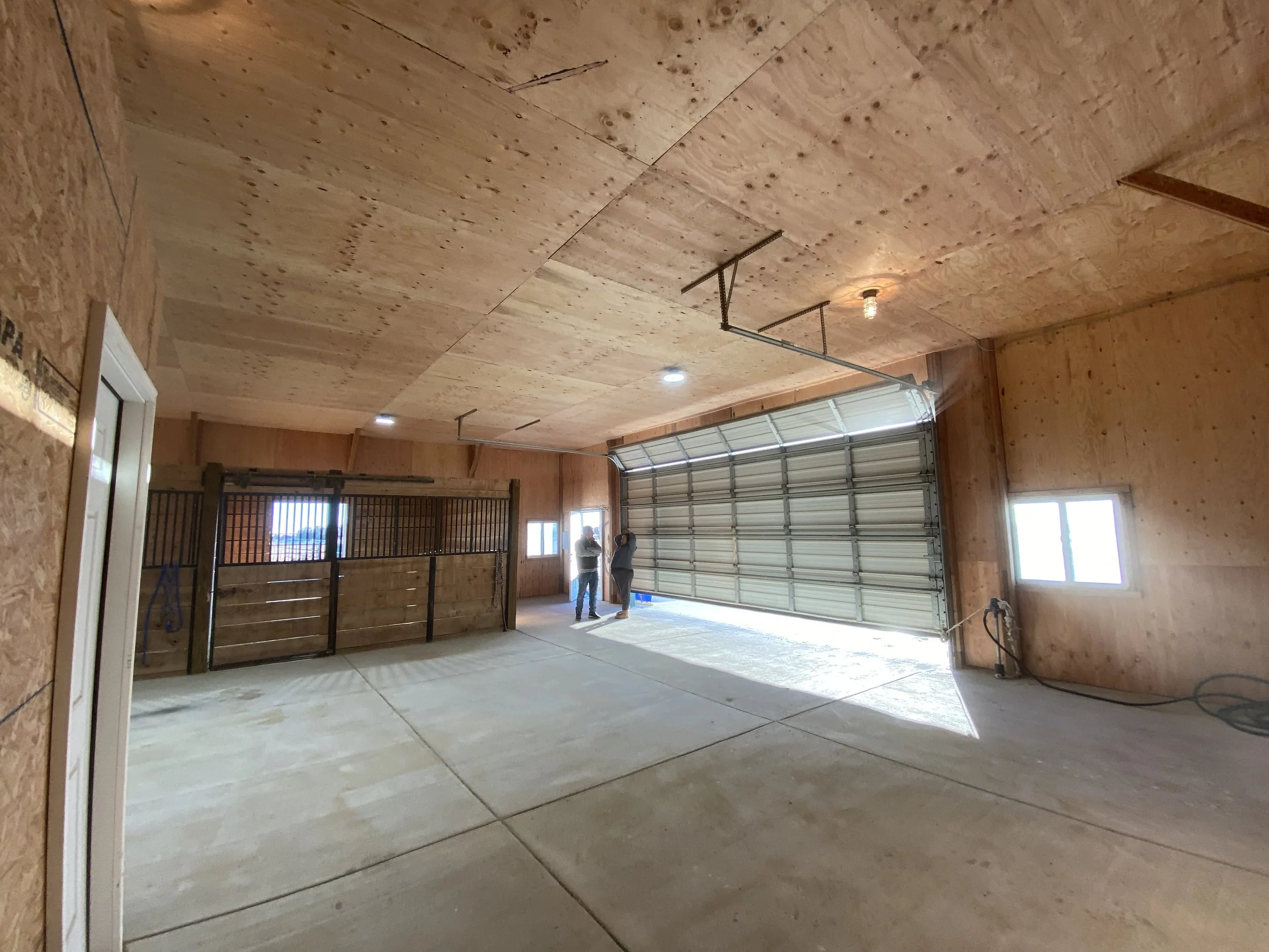 Interior view of a garage under construction with plywood walls and ceiling, concrete floor, and a partially open large garage door. Two people are standing near the garage door.