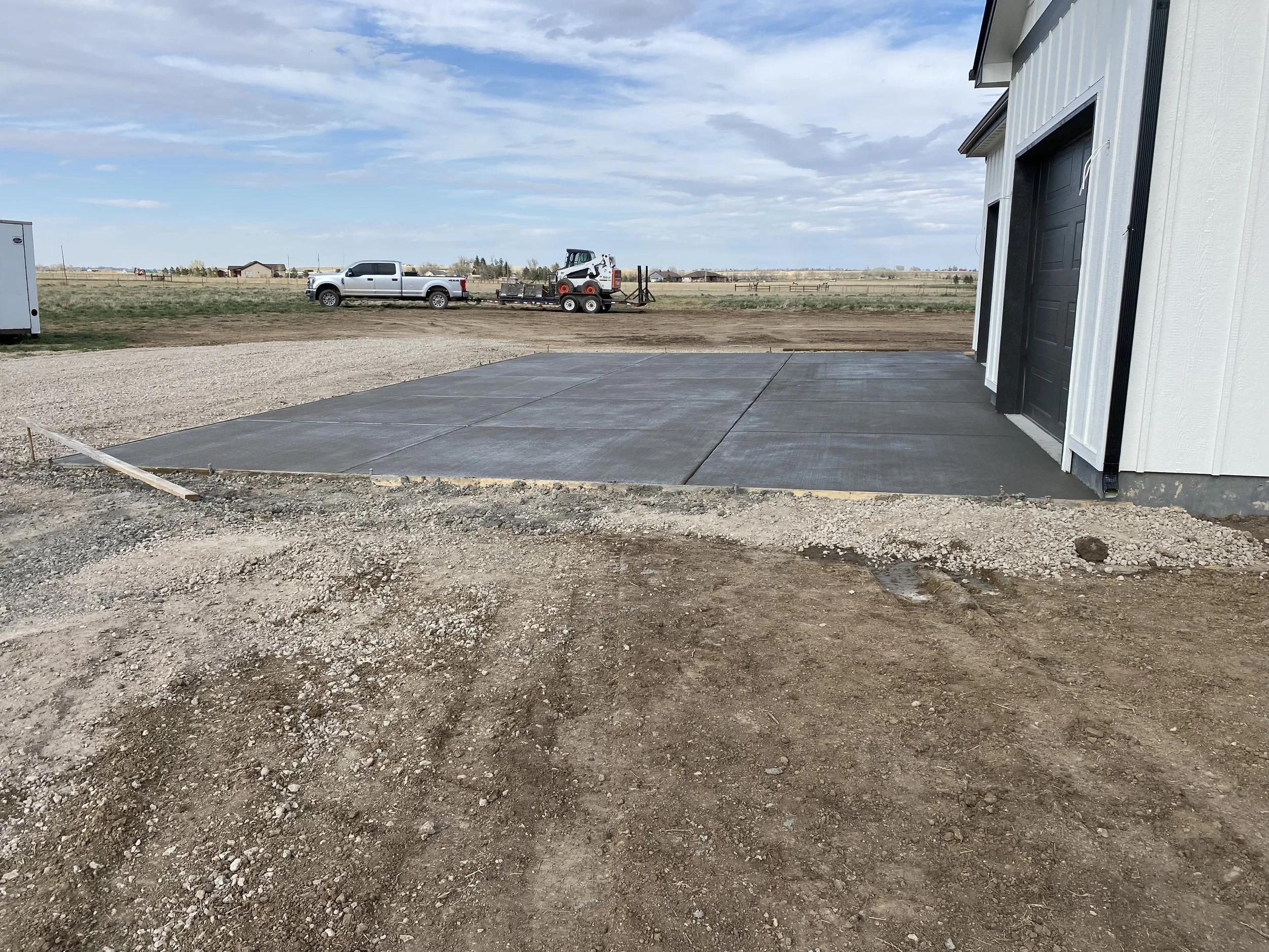 New concrete driveway next to a modern building with black garage doors, with a dirt and gravel landscape in the foreground and a flat open field in the background under a partly cloudy sky.