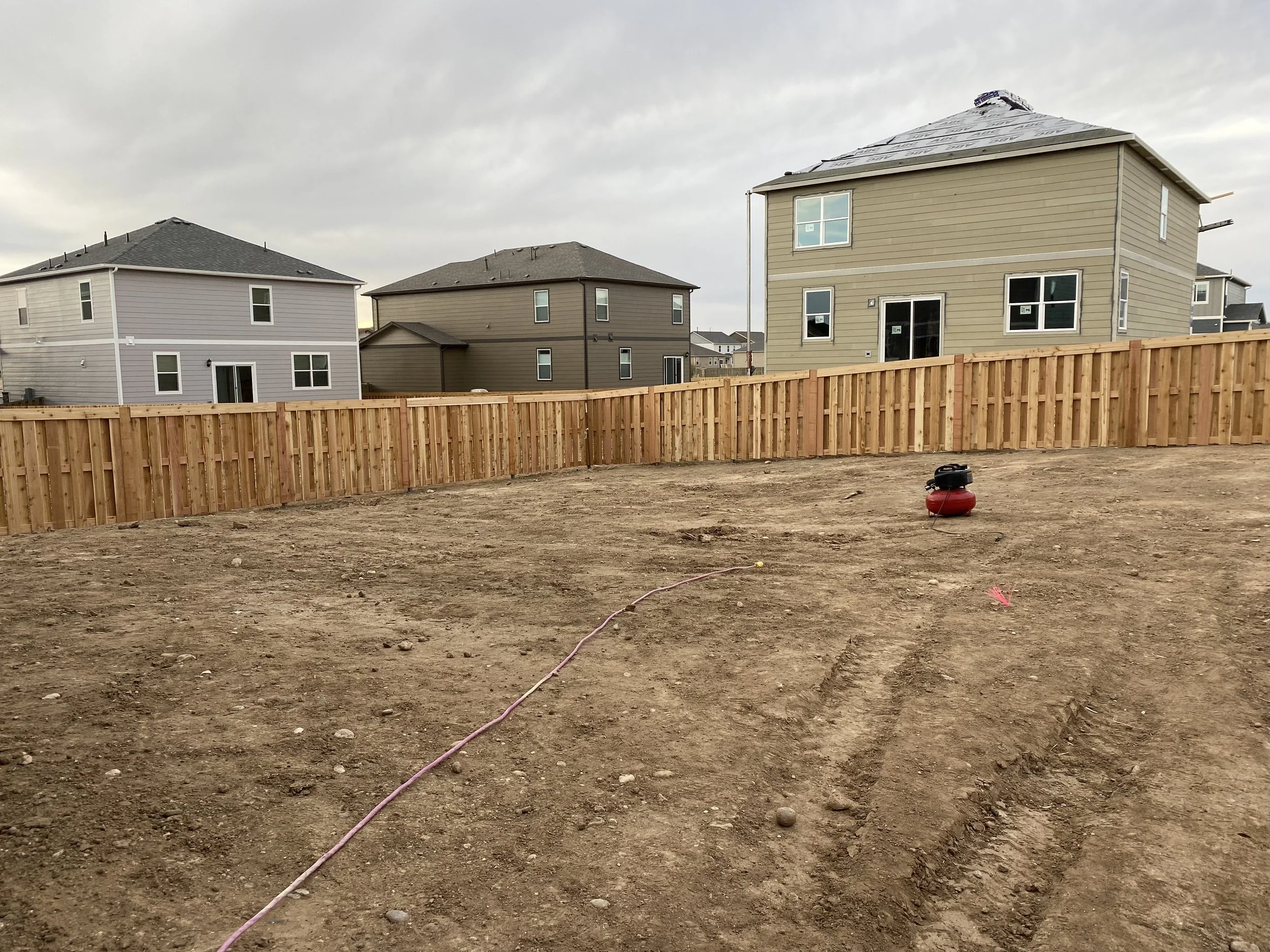 A backyard under construction with bare dirt, a pink string, and a red and black portable air compressor. Wooden fence surrounds the yard, with neighboring houses visible in the background under cloudy skies.