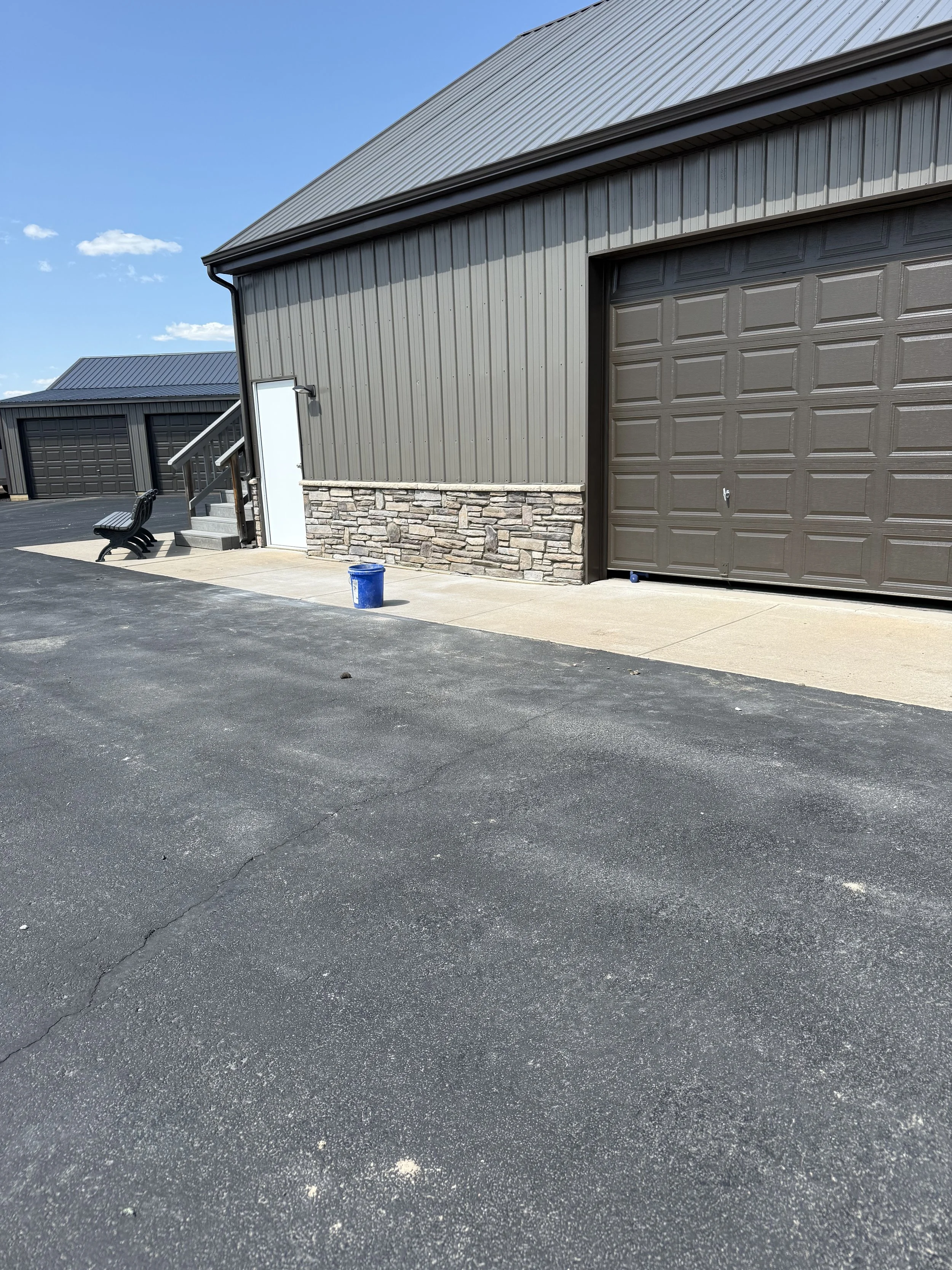 Exterior view of a large gray metal building with a stone lower section, a brown garage door, a small white door with stairs, a black bench, a blue bucket, and a paved lot under a clear blue sky.