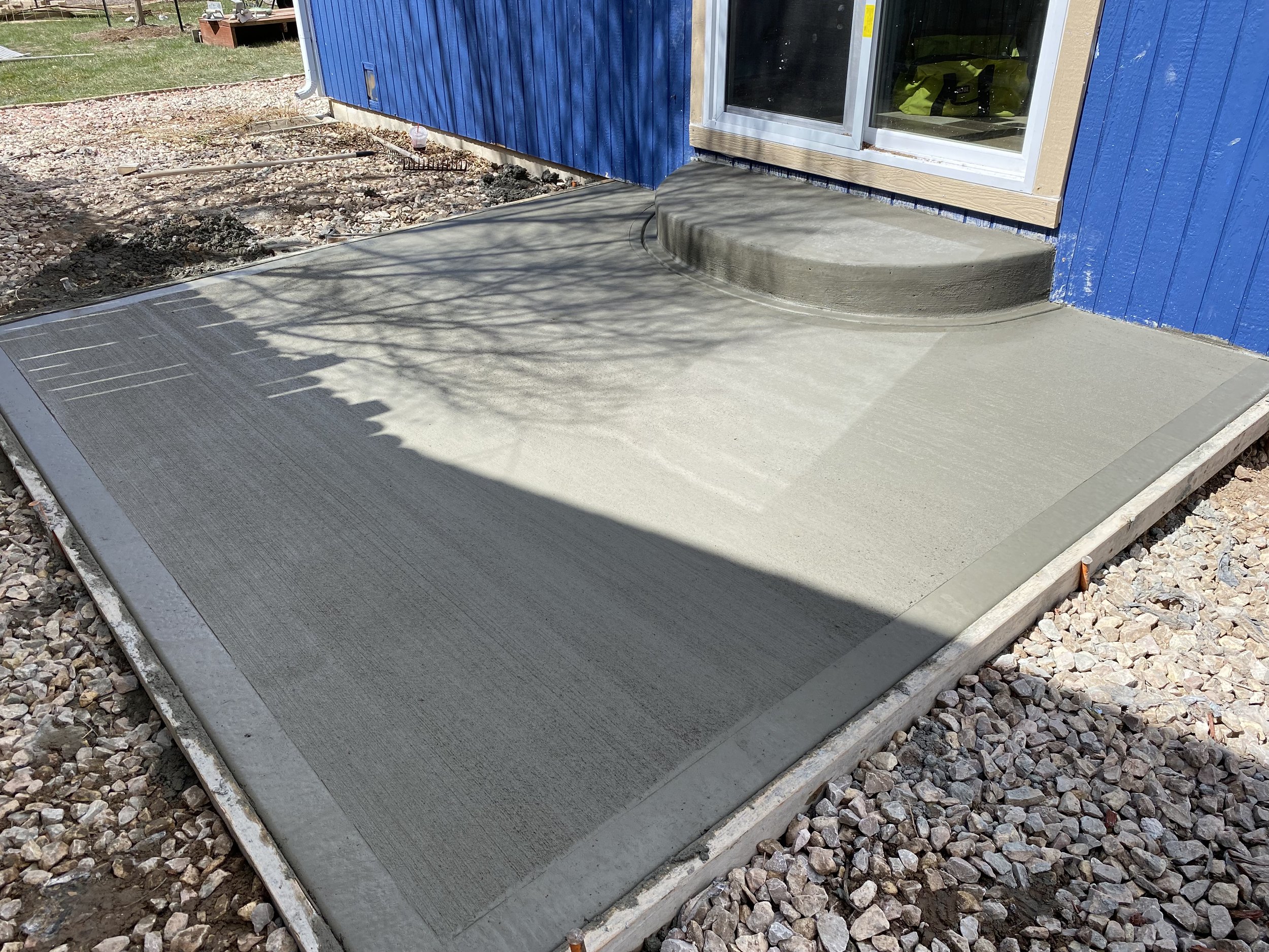 Freshly poured concrete porch at a house with blue siding, a sliding glass door, and gravel surround.