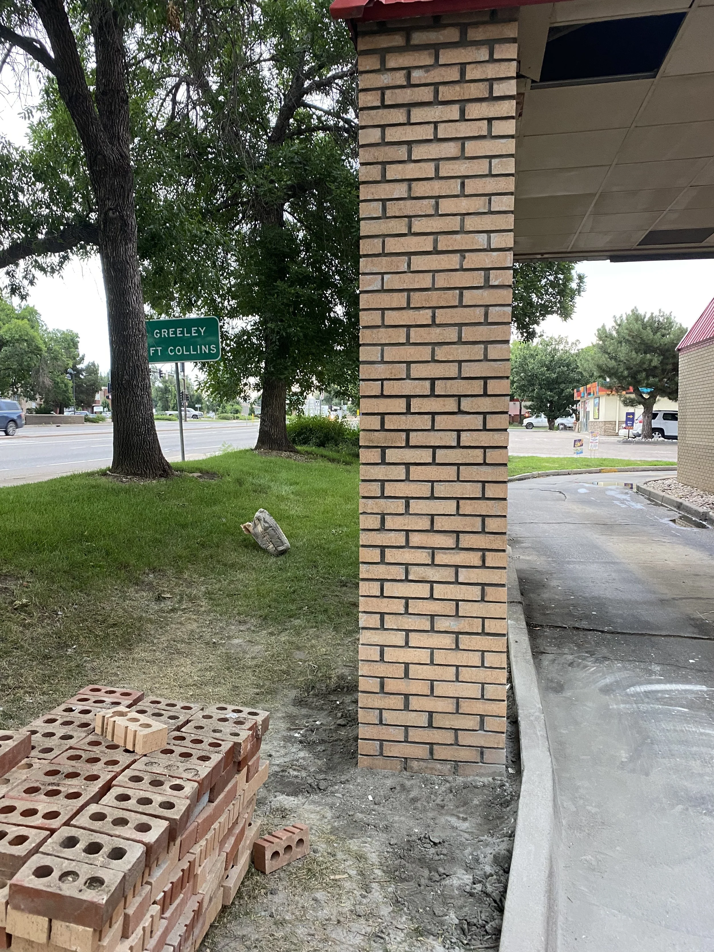 Construction site with stacks of red bricks and a brick wall under construction next to a sidewalk. Trees and a street with cars and a green street sign that reads "Greeley Ft Collins" are visible in the background.