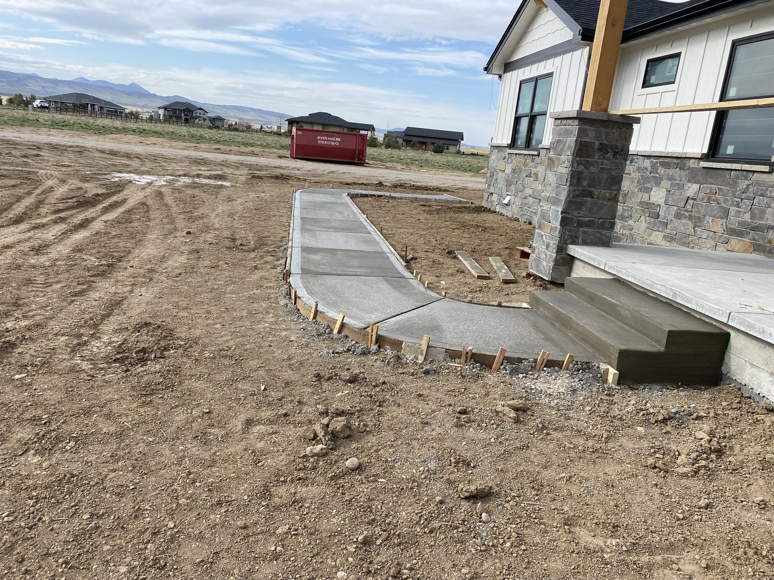 Construction site with newly poured concrete sidewalk leading to the front stairs of a house, with wooden forms remaining along the edge, dirt and construction materials on the ground, and a red dumpster in the background. The house has stone and siding exterior with large windows, and there are mountains visible in the distance under a partly cloudy sky.