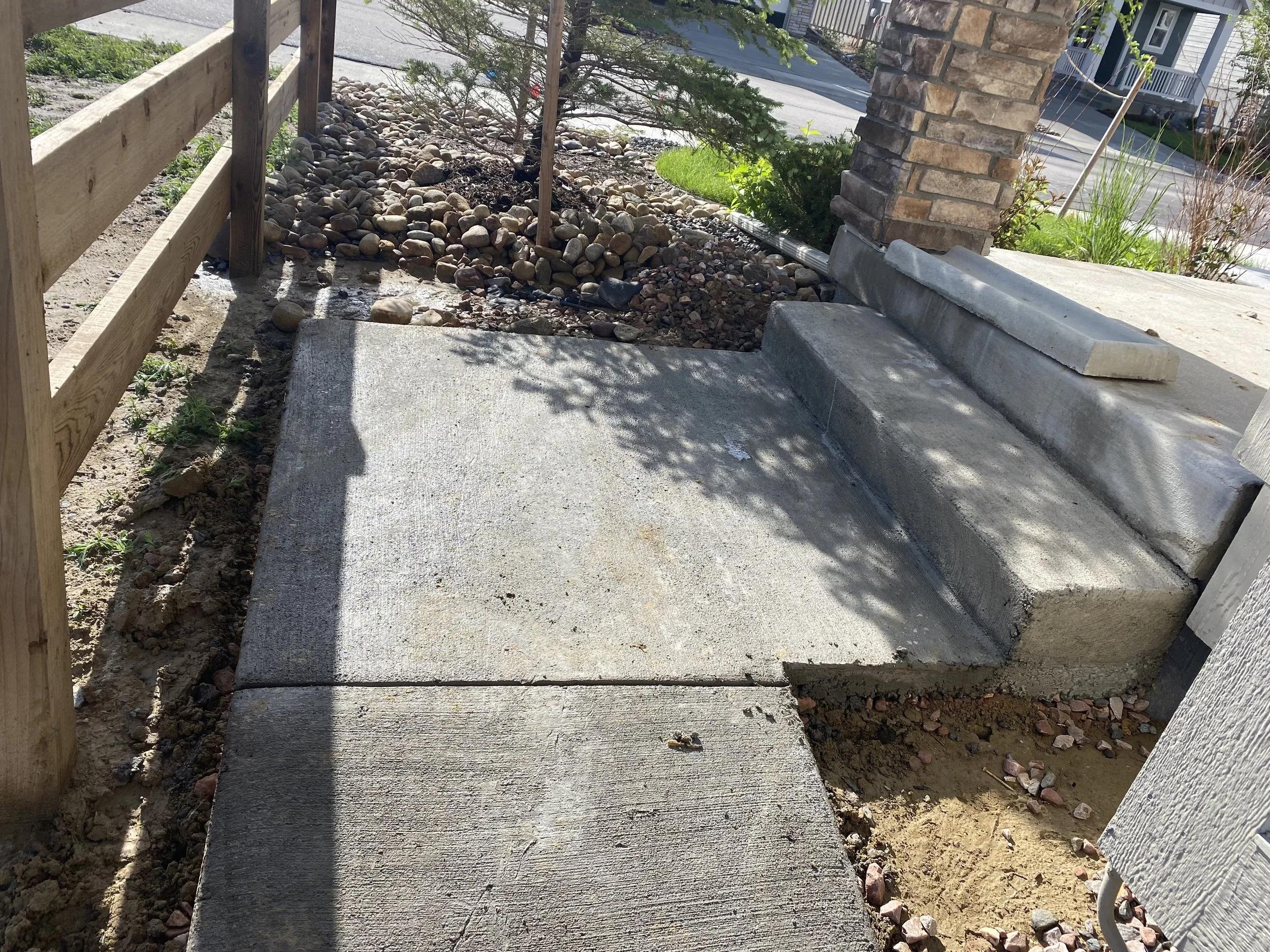 Concrete sidewalk and steps leading to a porch, with a brick pillar and a wooden fence on the left, and landscaping with rocks and plants in the background.