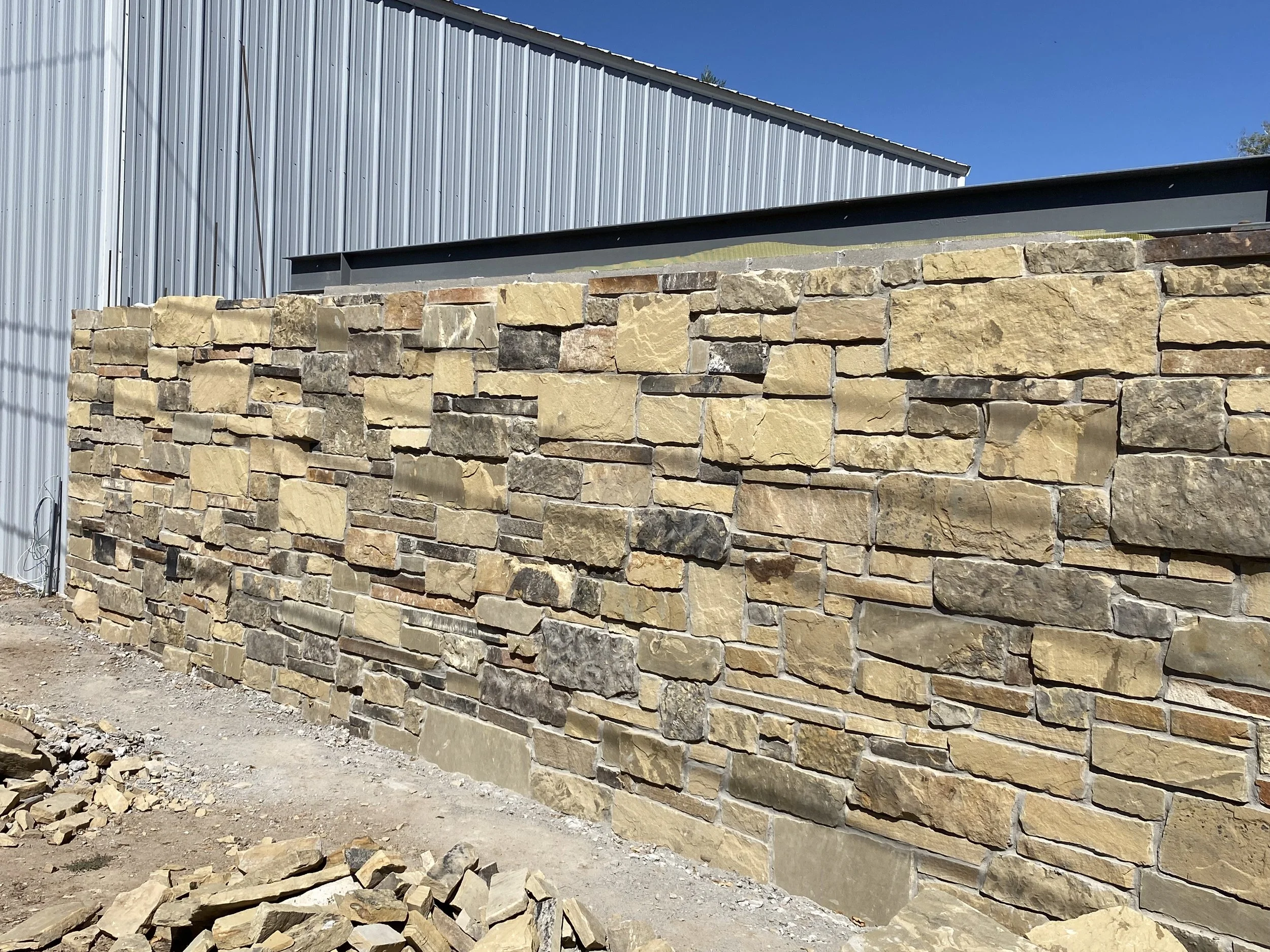 A stone wall under construction with a metal building in the background and a clear blue sky.