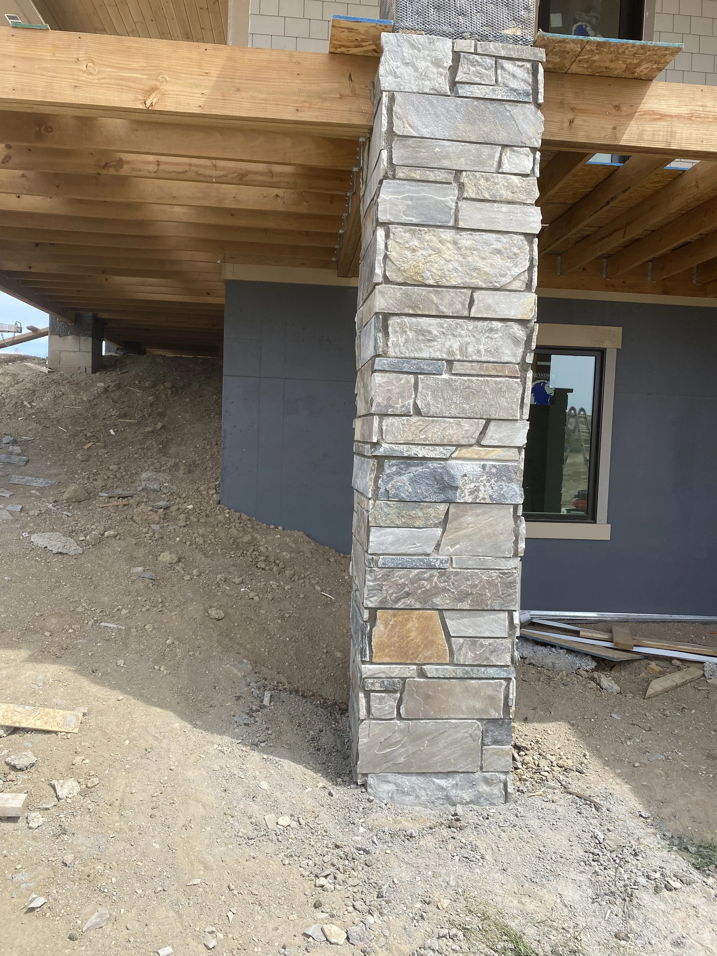 Close-up of a stone pillar supporting a wooden deck at a construction site, with dirt and construction materials on the ground.
