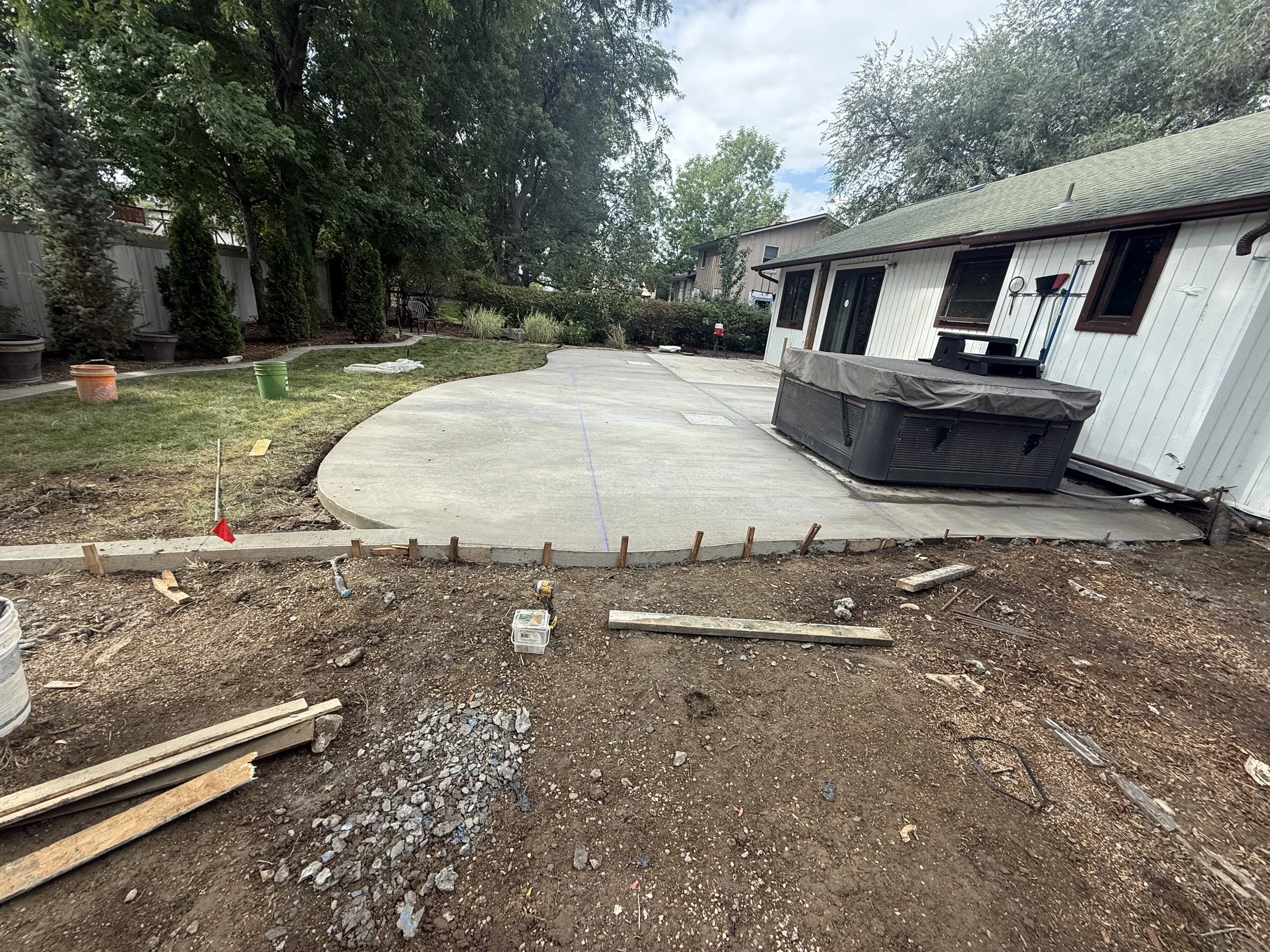 Backyard patio under construction with a freshly poured concrete slab, surrounded by dirt and construction materials, on the side of a white house with green roof.