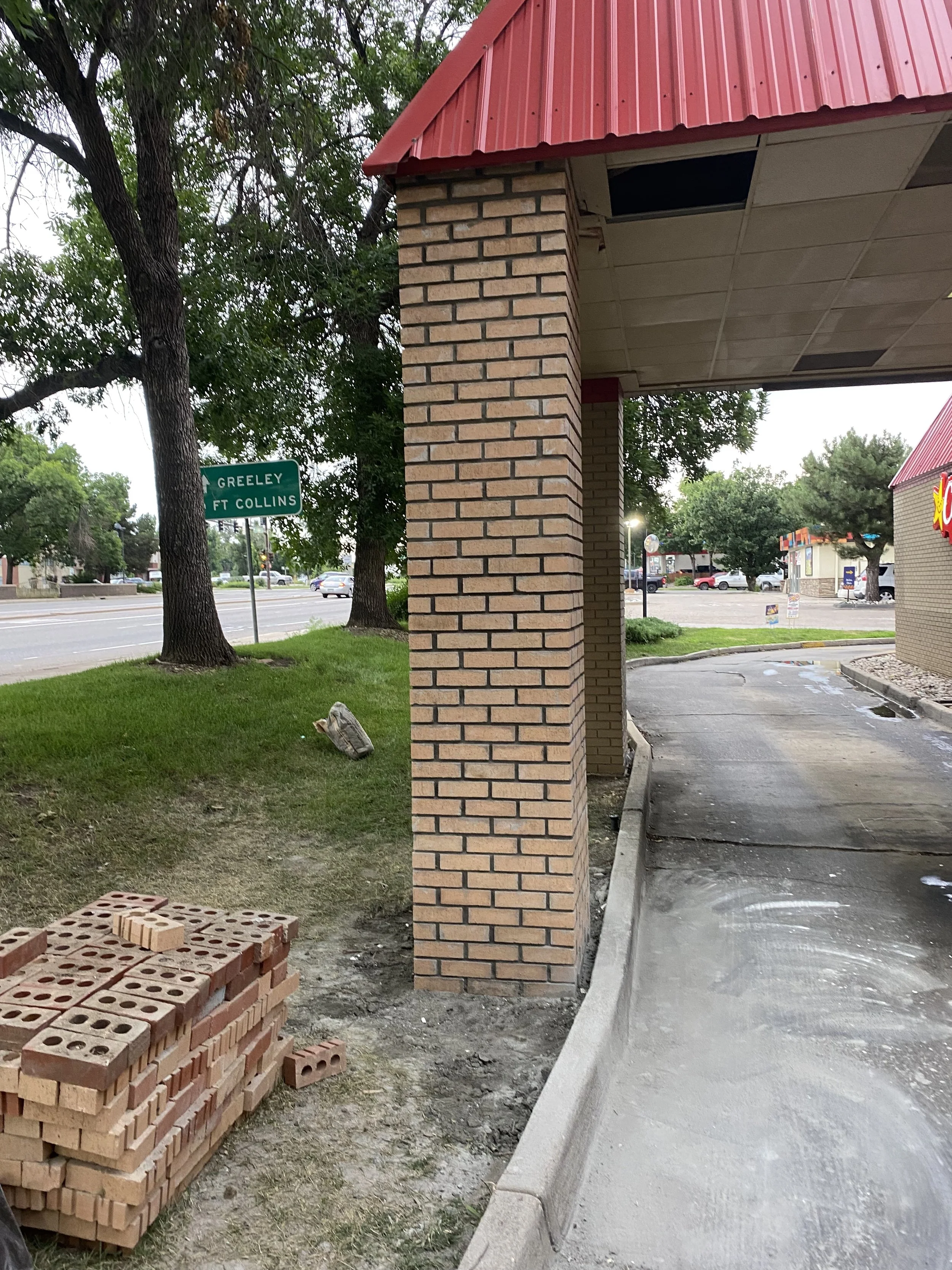A sidewalk and part of a building with a brick column and a red metal roof, trees in the background, and a street sign indicating directions to Greeley and Ft Collins across a street.