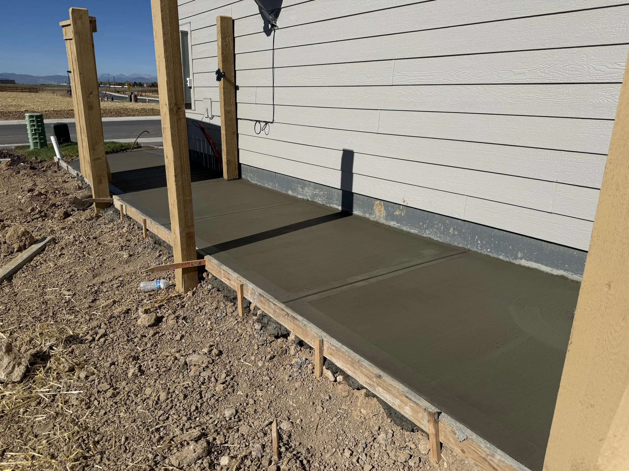 Freshly poured concrete walkway in front of a house under construction, with wooden stakes supporting formwork, on a sunny day.