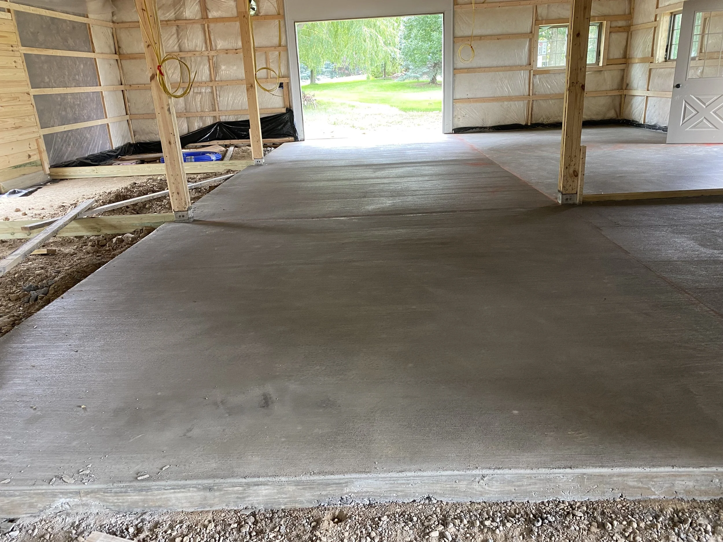 View inside a house under construction showing new concrete flooring, exposed wooden framing, and an open garage door leading outside to a green yard with trees.