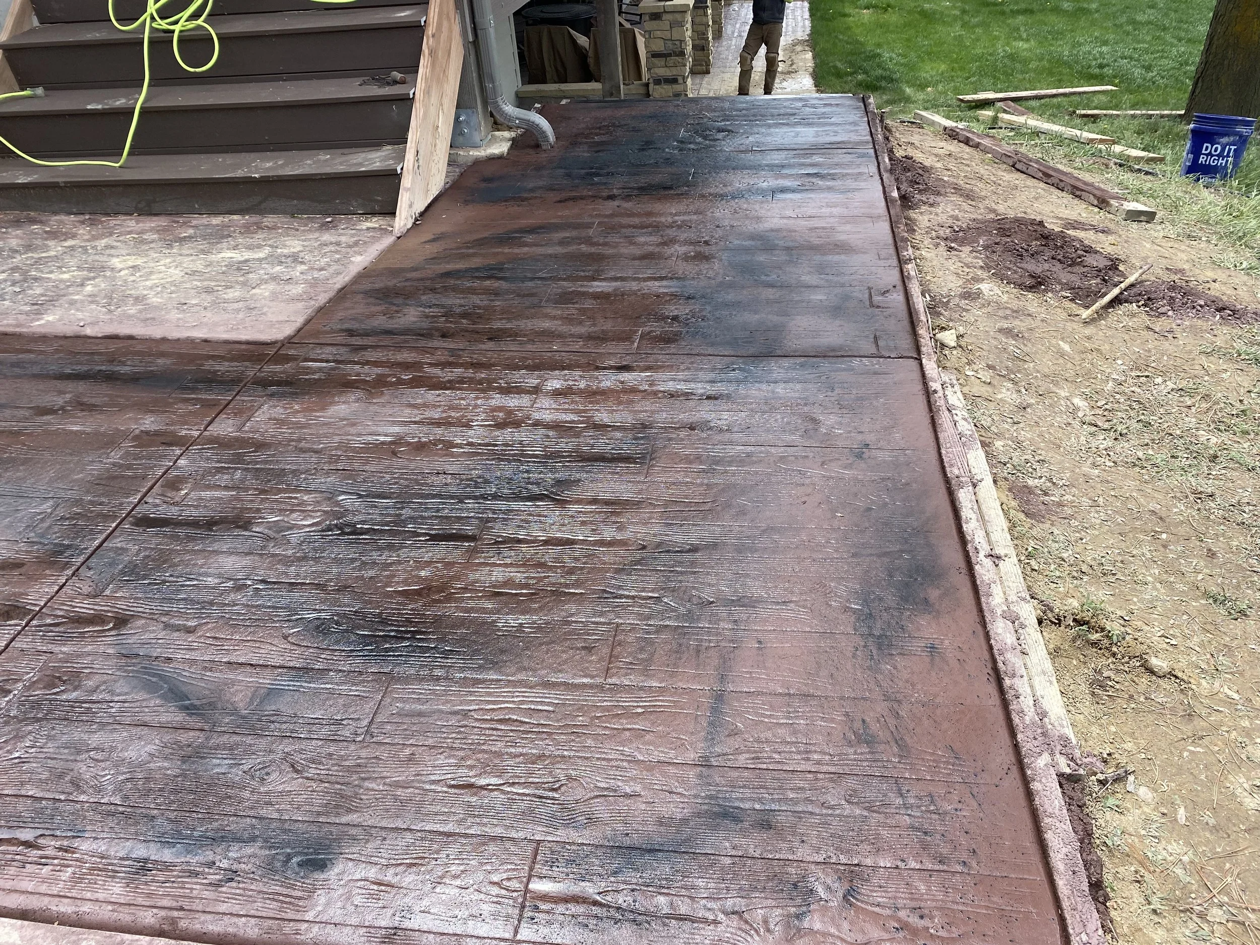 A freshly stained wooden deck outside a house, with a section of it still unpainted or lightly stained. The house has siding, and there are garden tools and construction materials visible nearby.