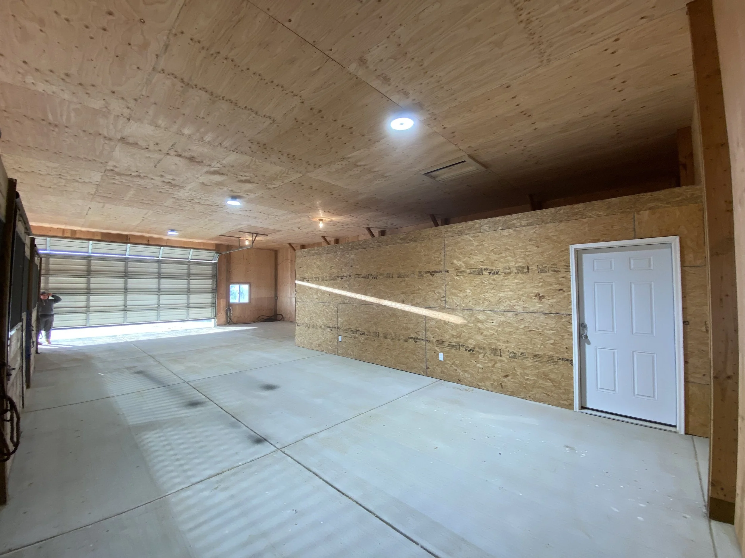 An unfinished garage with a concrete floor, wooden walls and ceiling, a large metal garage door, a small window, and a closed white door.