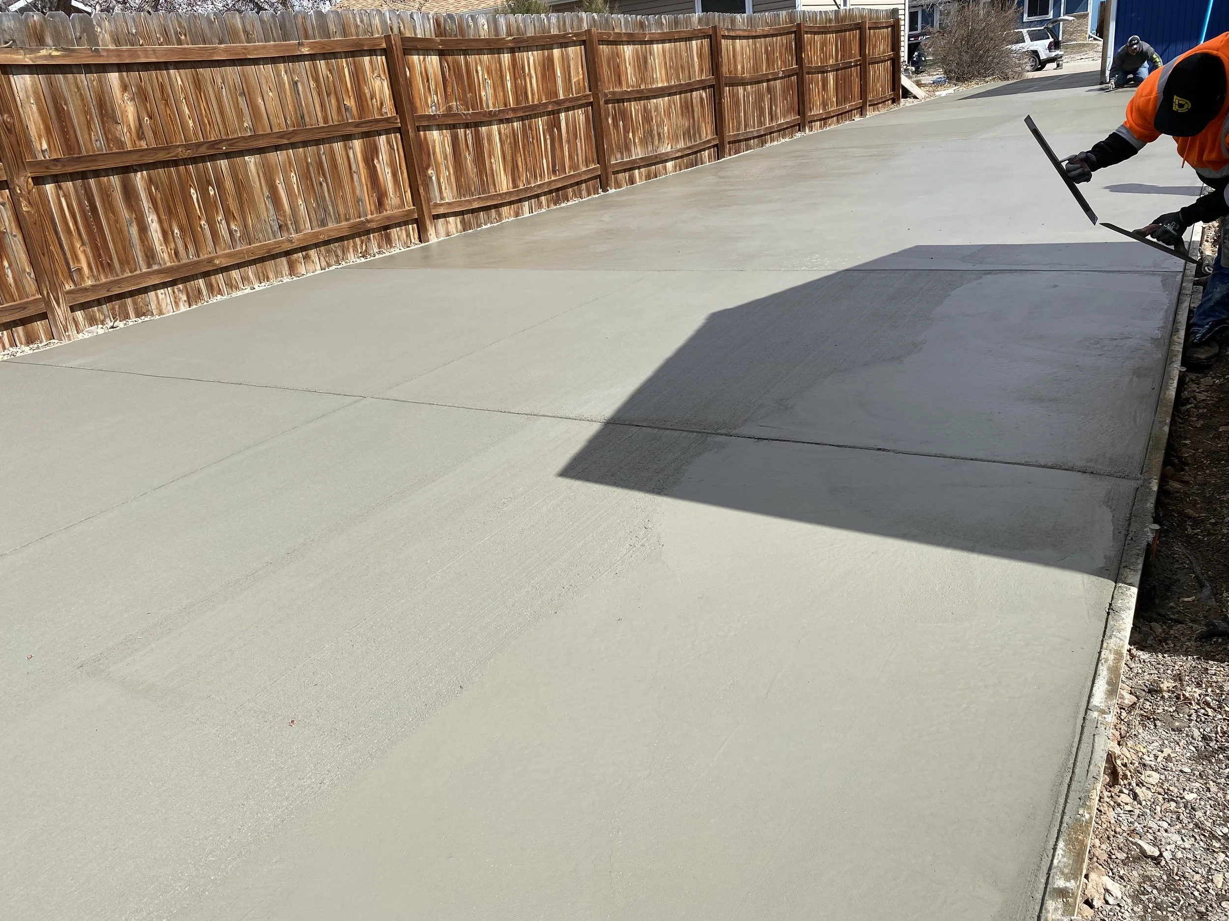 Workers are finishing pouring a concrete driveway, smoothing the surface with hand tools. A wooden fence runs along the left, with houses and parked cars in the background.