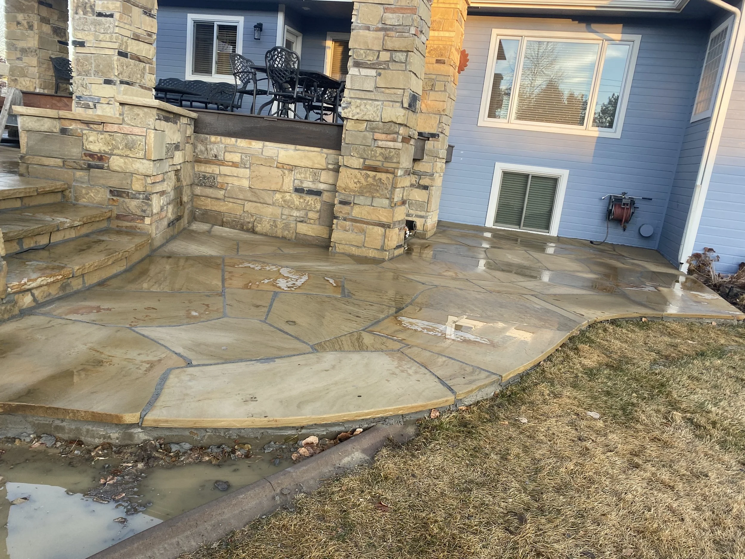 A stone outdoor patio with curved steps leading to a raised deck area with patio furniture, and a house with blue siding and white windows in the background.