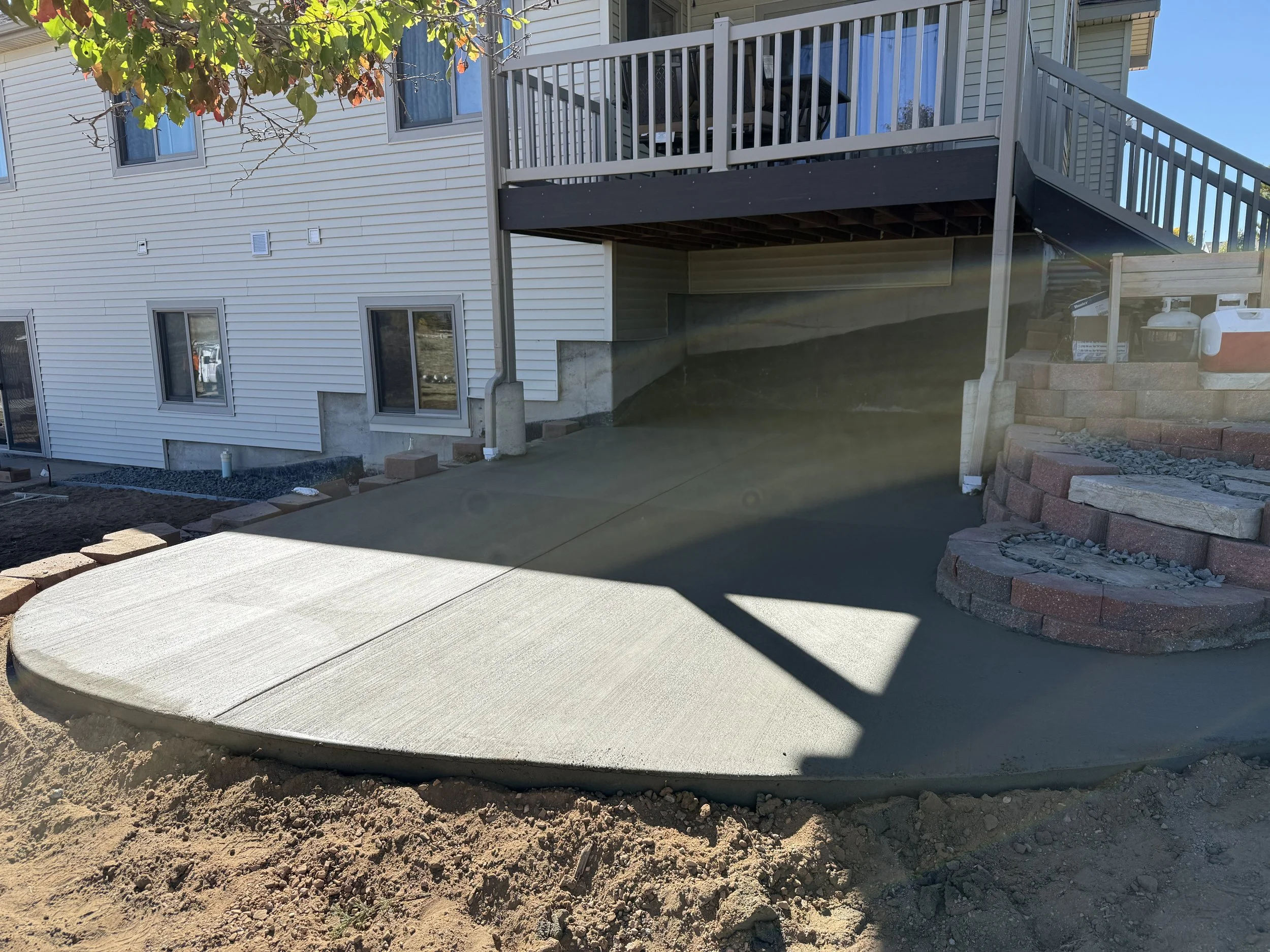 Newly poured concrete patio with a curved edge outside a house with a raised deck and stairs; construction materials and tools visible on the side.
