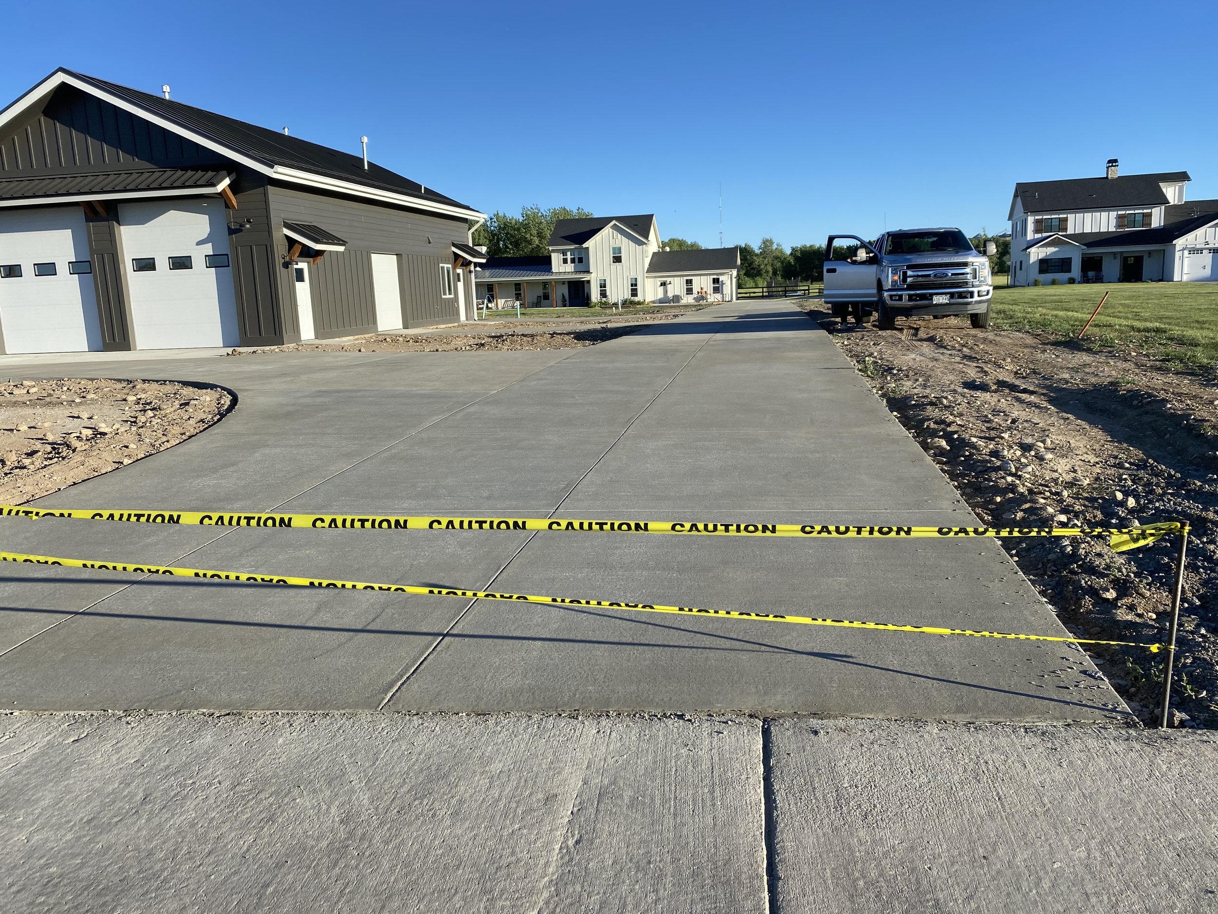 Newly paved concrete driveway with caution tape across it, leading to a residential area with houses, a truck, and a clear blue sky.